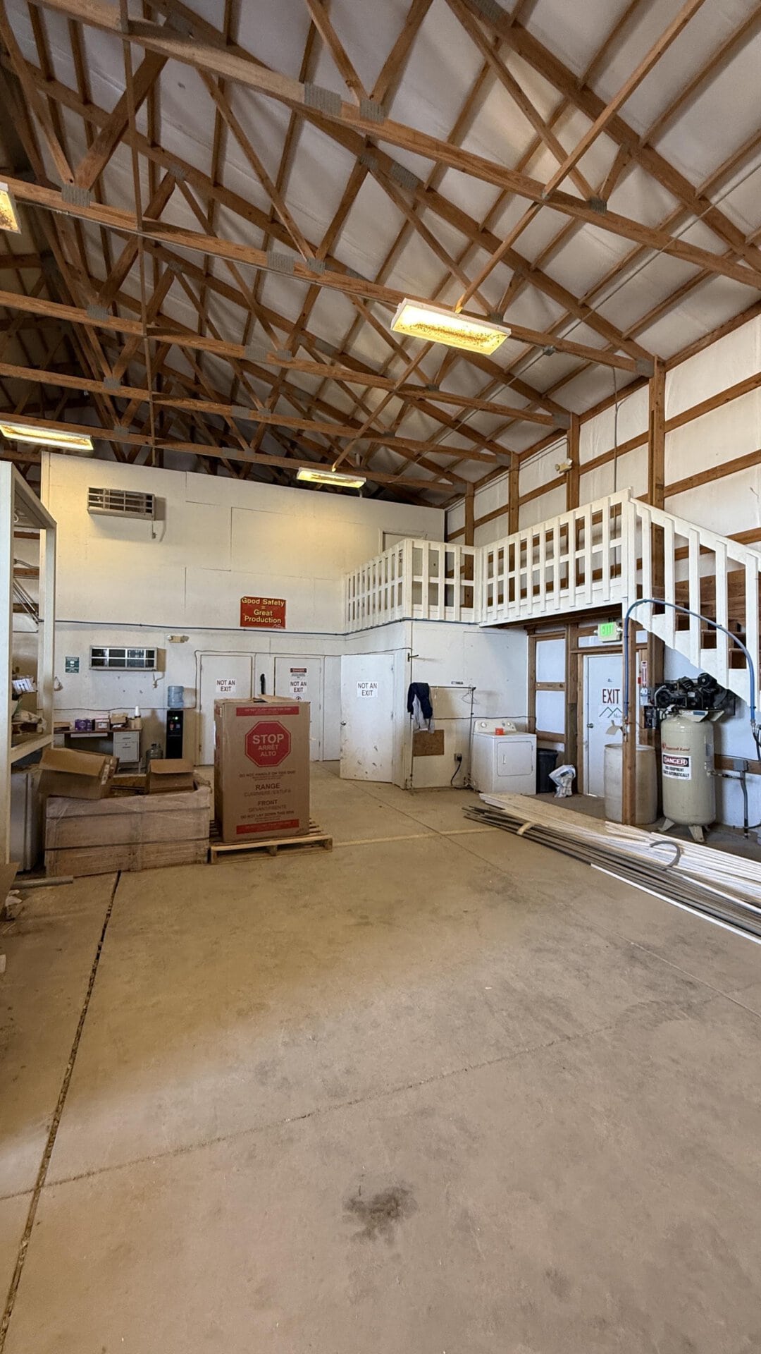 Industrial workshop interior with exposed wooden trusses, mezzanine railing, and overhead fluorescent lights.