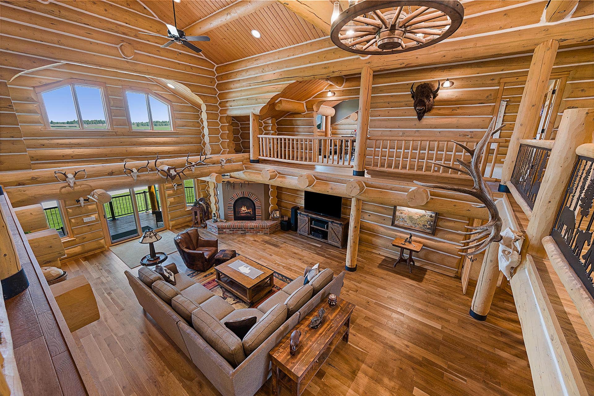 Open-concept log cabin living room with vaulted ceiling, large windows, and a brick fireplace surrounded by logs and antler decor on the walls.