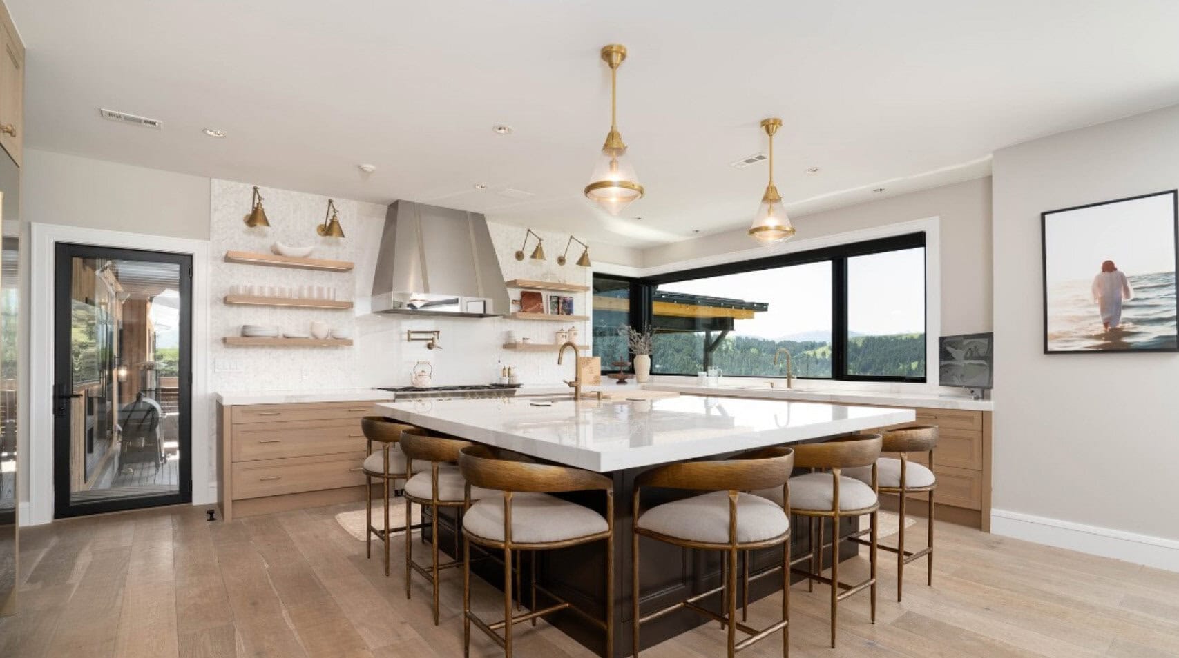 Spacious modern kitchen with a large white island, wooden stools, and open shelves against a bright wall with a window view of trees outside.