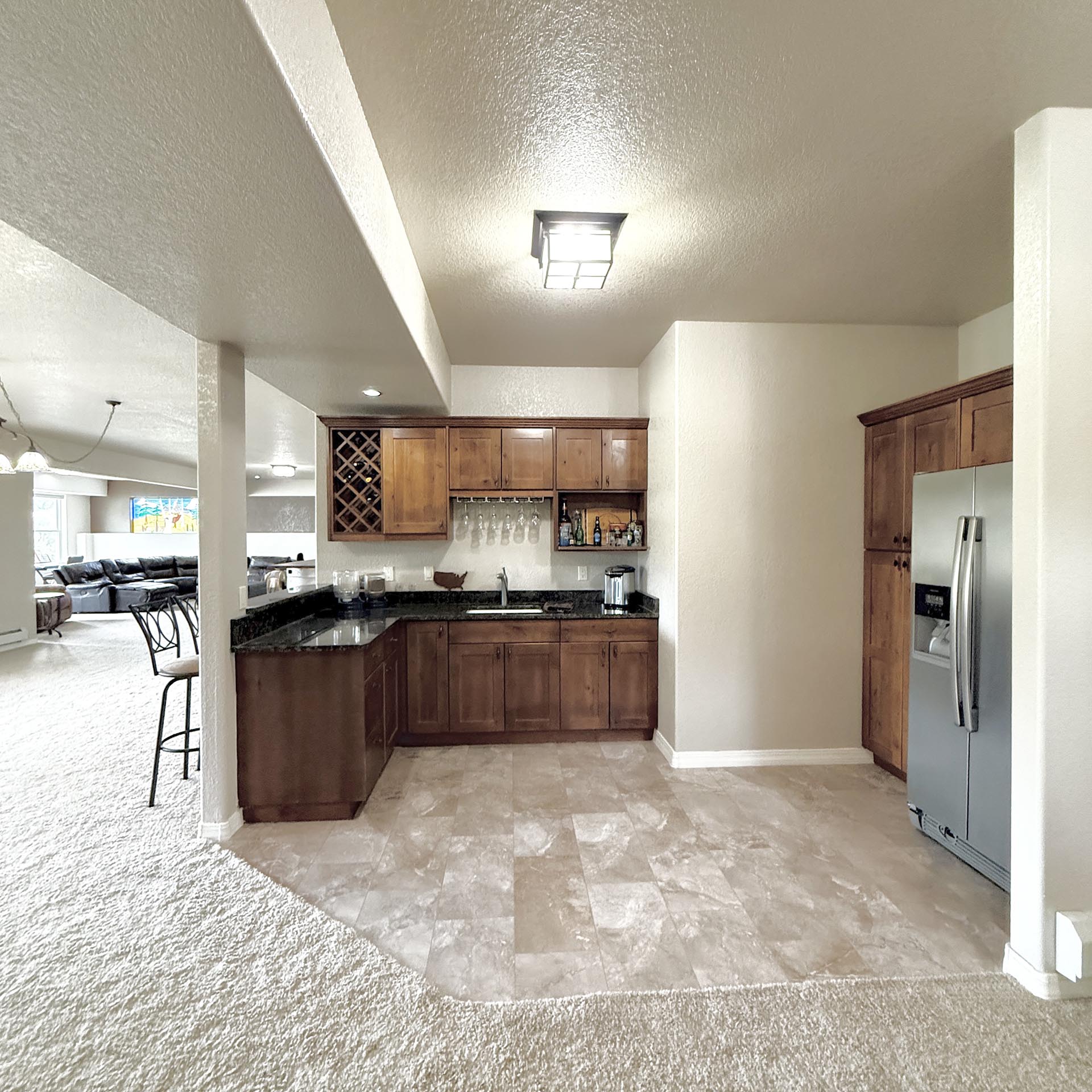 Open-concept kitchen with dark wood cabinets, wine rack, hanging glassware, and a stainless steel refrigerator; living area visible in the background.