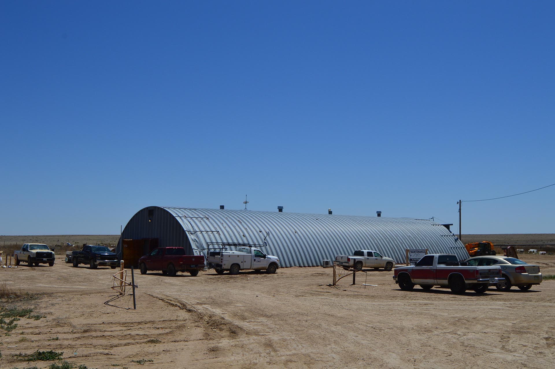 Large corrugated metal hangar in a dusty desert lot with several pickup trucks parked in front and a maintenance sign nearby on a bright day.