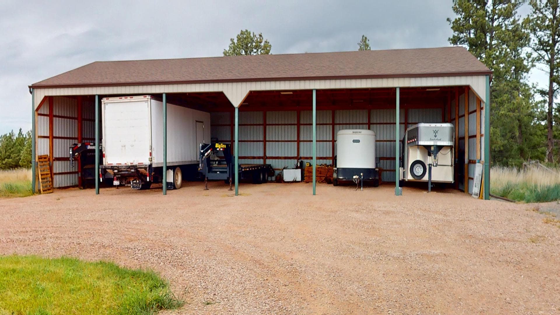 Open-sided metal storage structure on a gravel lot housing a box truck, a trailer, and other equipment with a white trailer and a fuel tank visible, set against a rural backdrop of trees and grass.