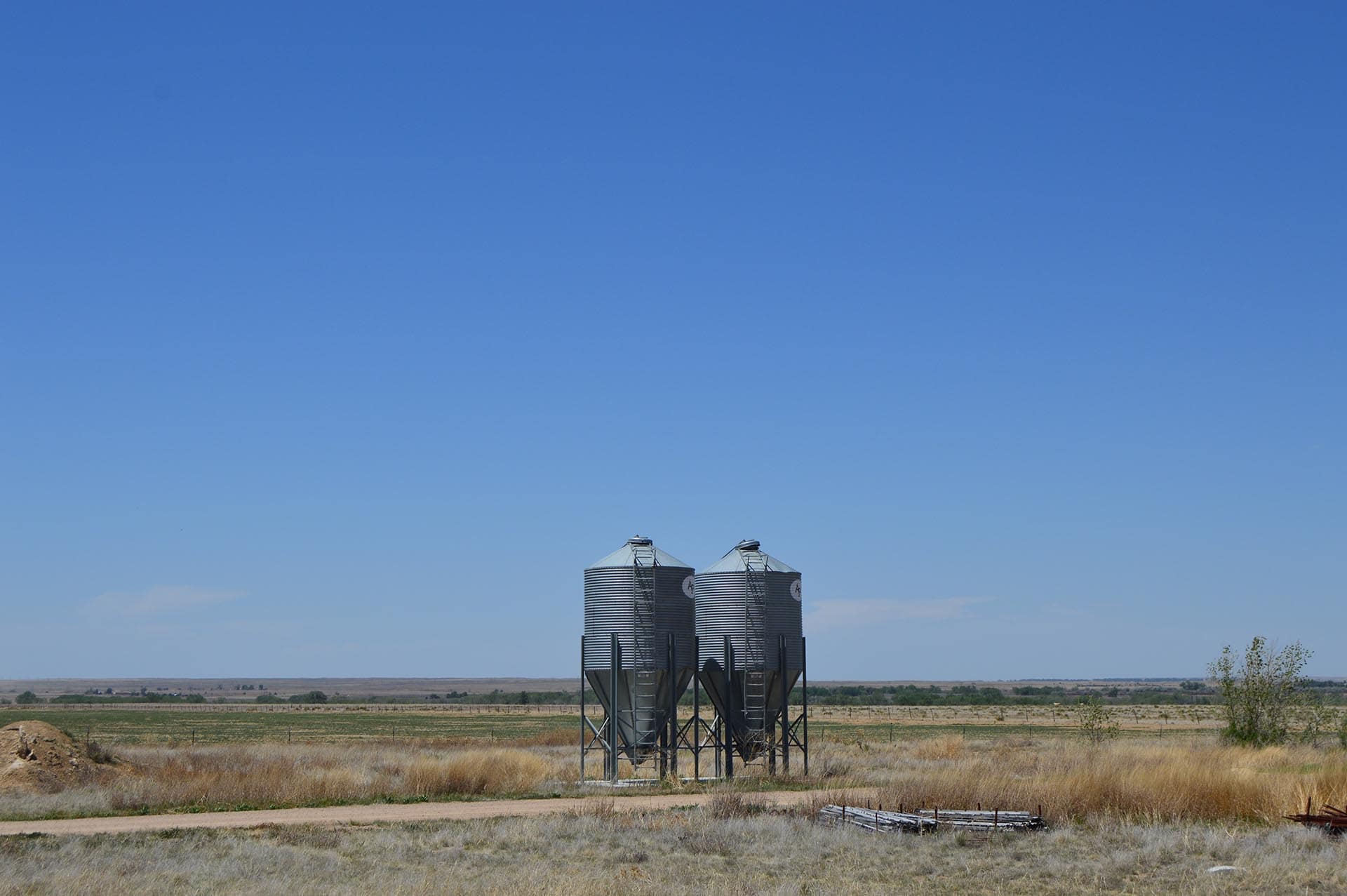 Two tall metal grain silos on a raised frame in a dry rural field under a clear blue sky.