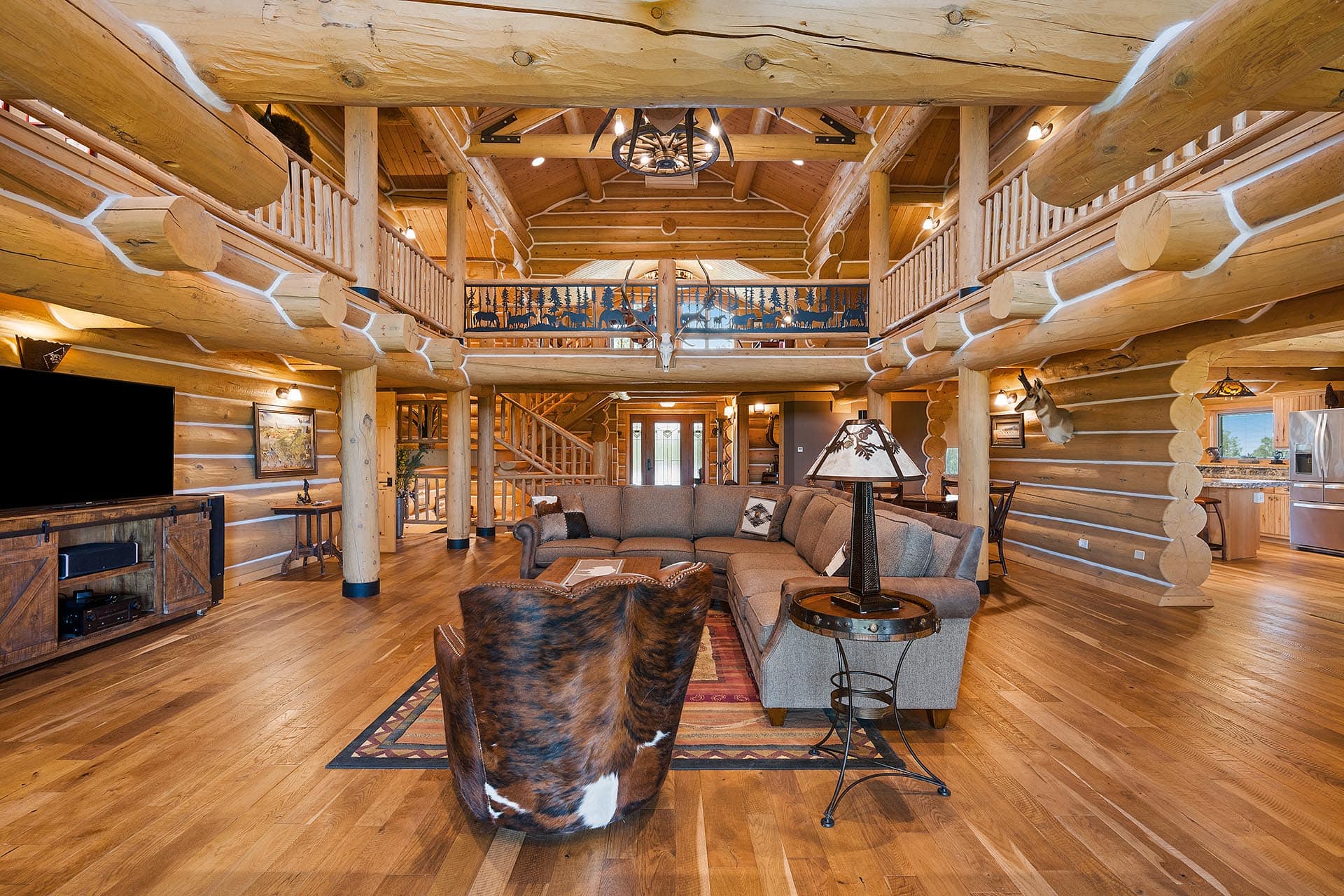 Spacious log cabin living room with exposed timber beams, large sectional sofa, cowhide chair, and kitchen visible in the background.