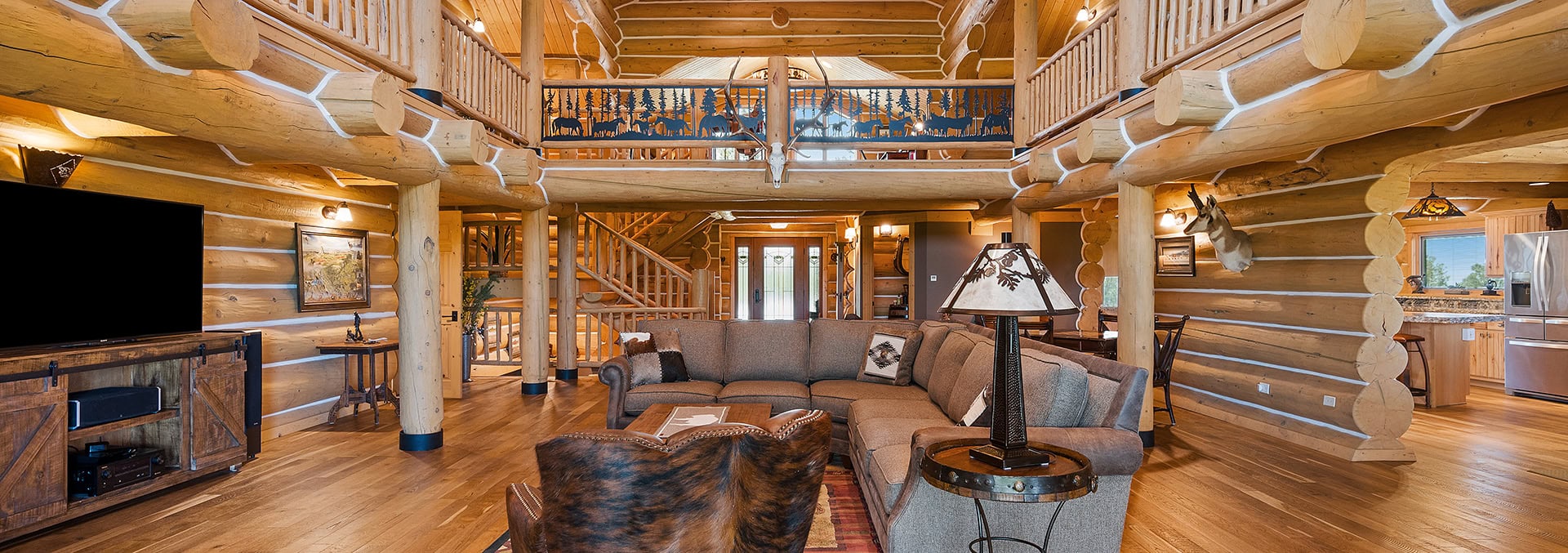 Rustic log cabin living room with an L-shaped gray sectional, cowhide chair, and a round metal side table under a log-beam ceiling.