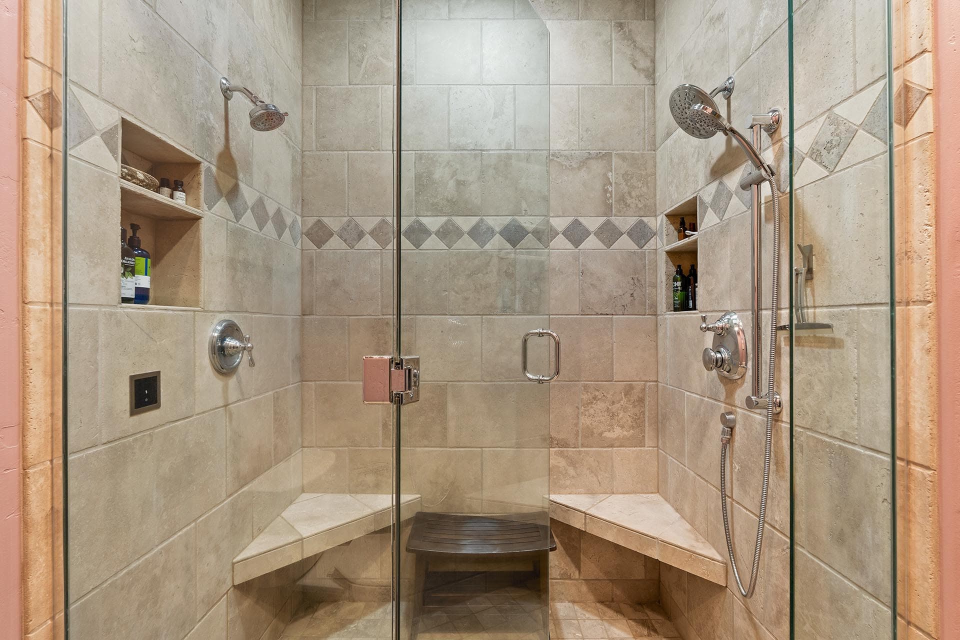 Walk-in tiled shower with glass doors, two wall-mounted showerheads, a built-in corner bench, and recessed shelves holding bath products.