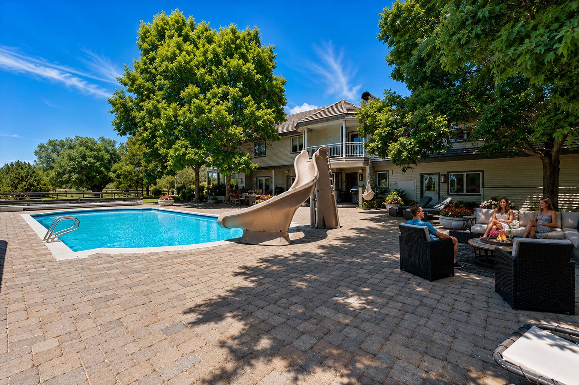 Poolside backyard with a beige slide, lounge seating, and people relaxing around a fire pit by a two-story house under trees.