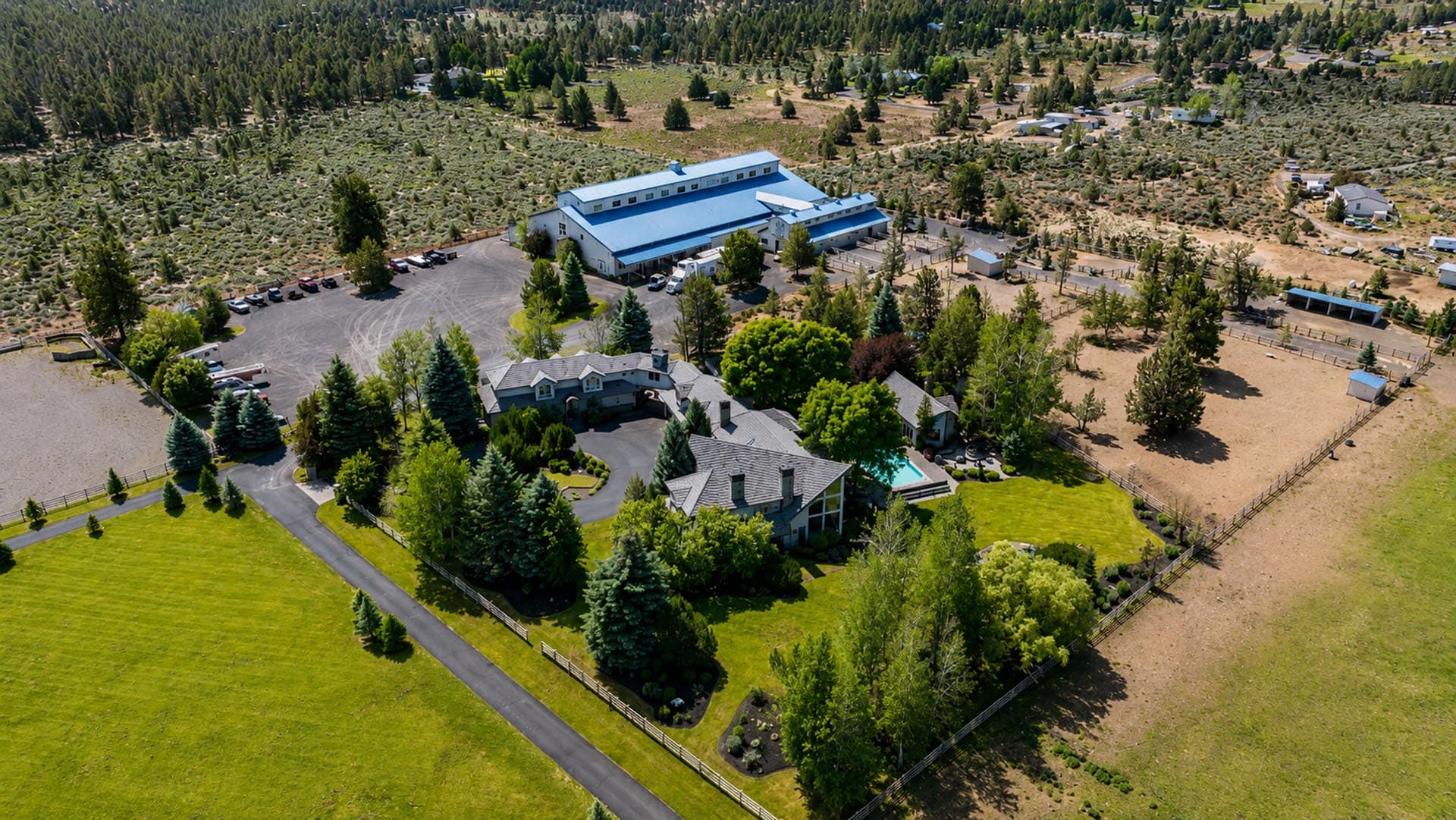 Aerial view of a large estate with a white house, blue-roofed buildings, a swimming pool, and extensive manicured lawns surrounded by trees and open land.