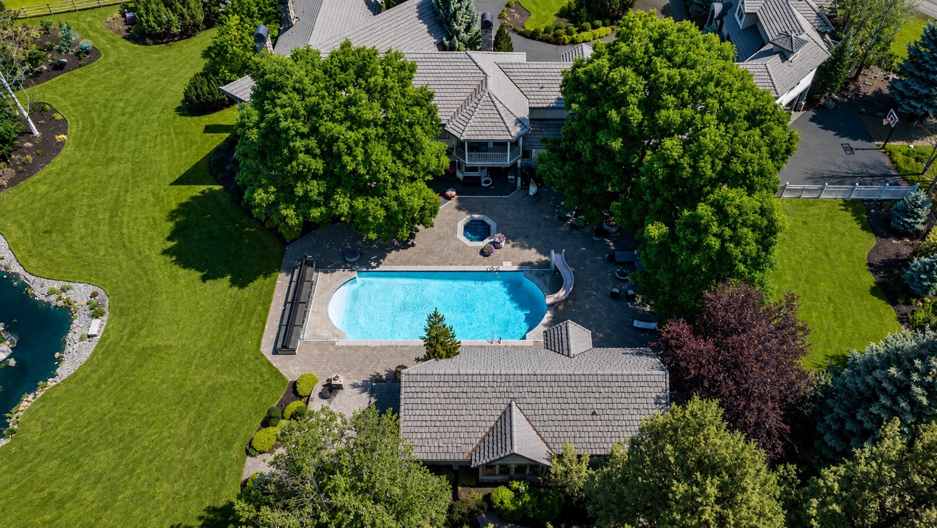 Aerial view of a large backyard with a rectangular pool, a hot tub, and a circular spa area surrounded by a paved patio and green lawn.