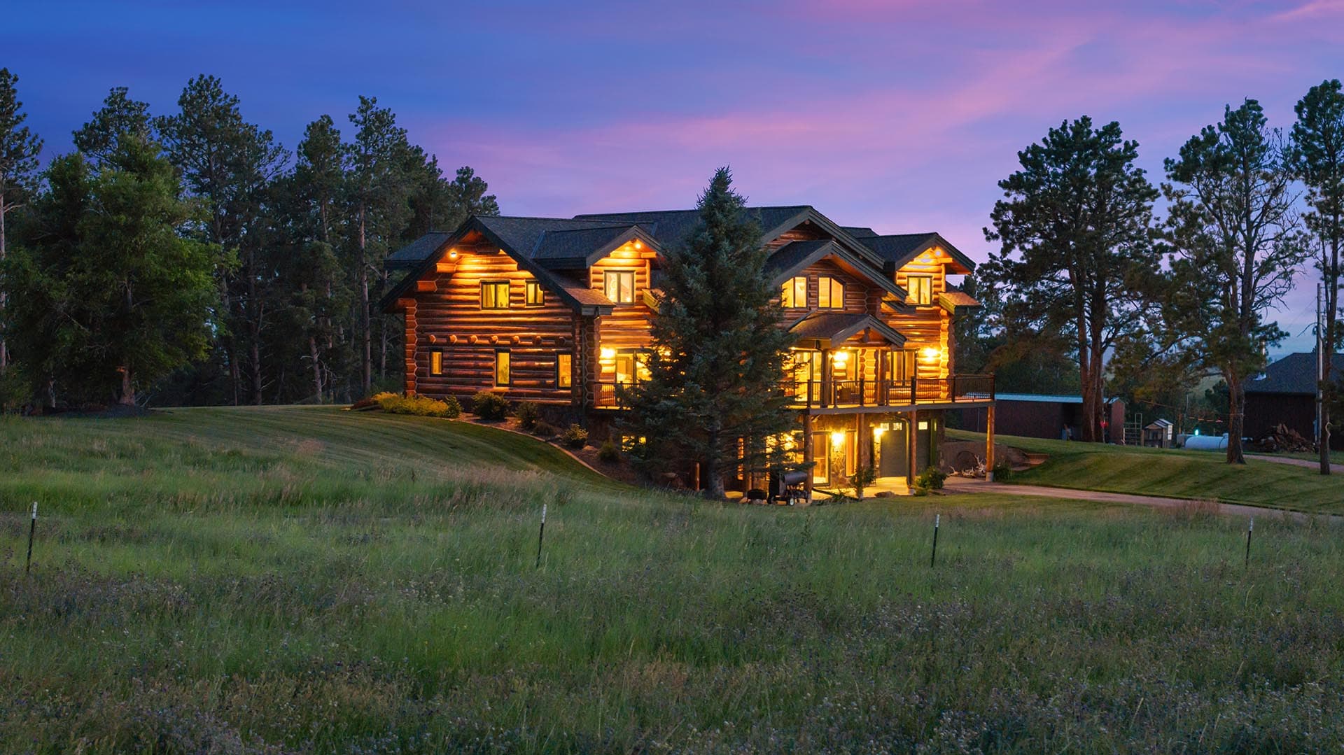 A two-story log cabin with warm interior lights, on a grassy slope surrounded by trees at dusk.