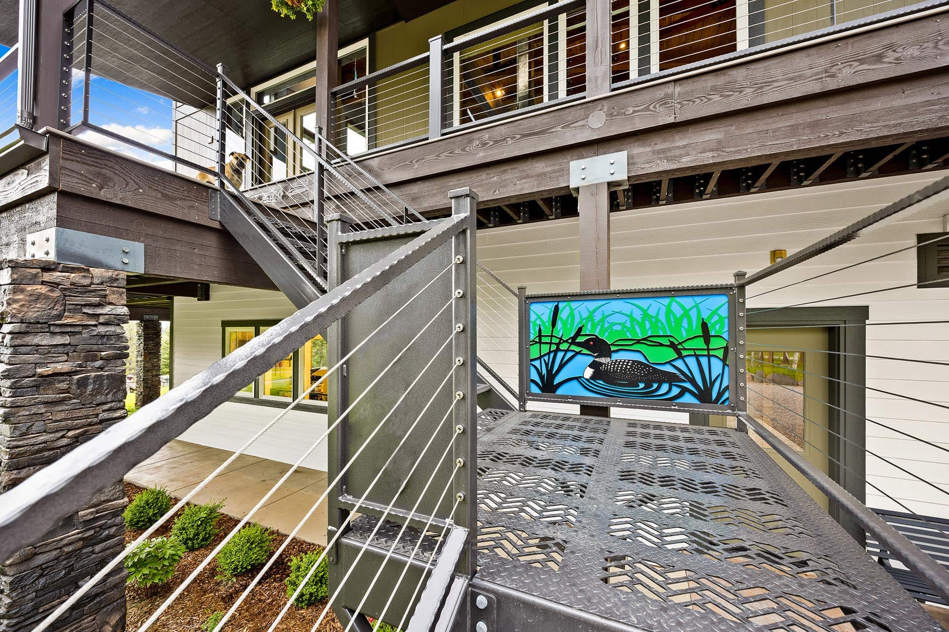 Exterior metal staircase with cable railing leading to a wooden deck; a colorful mural panel shows a waterfowl in a marsh.