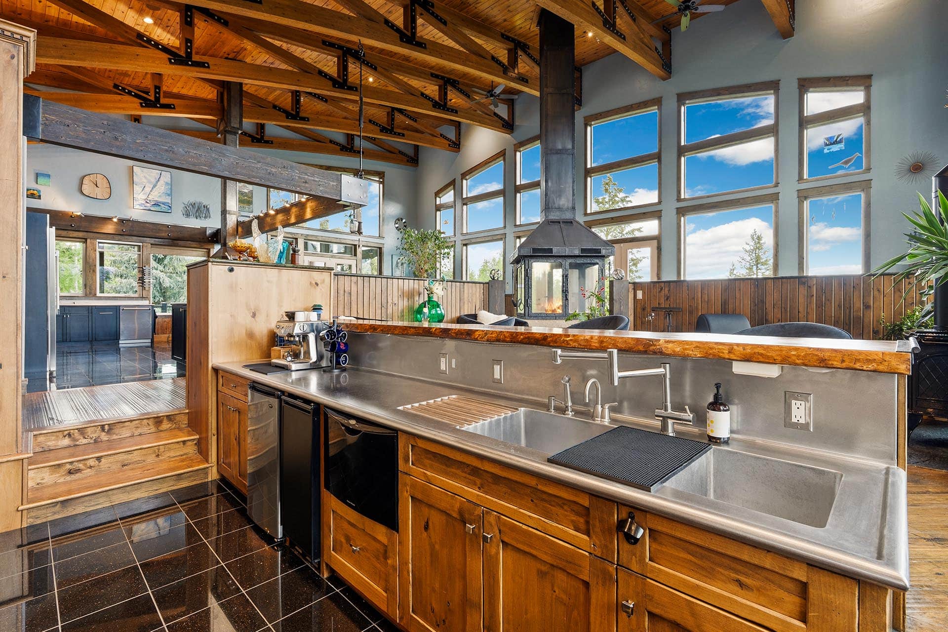 Open-concept kitchen with wood cabinetry, stainless steel counters, and a tall metal chimney fireplace at the center under a vaulted beam ceiling.