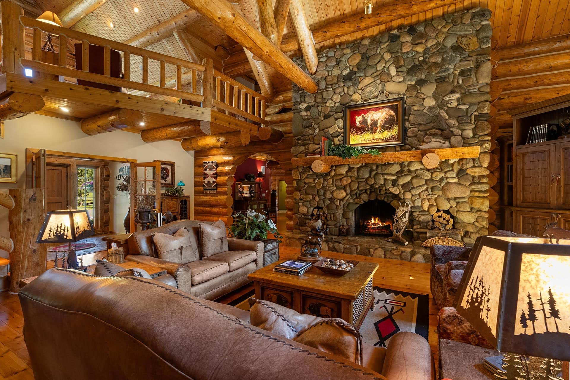 Cozy log cabin living room with a towering stone fireplace, wooden beams, and leather sofas arranged around a wooden coffee table.