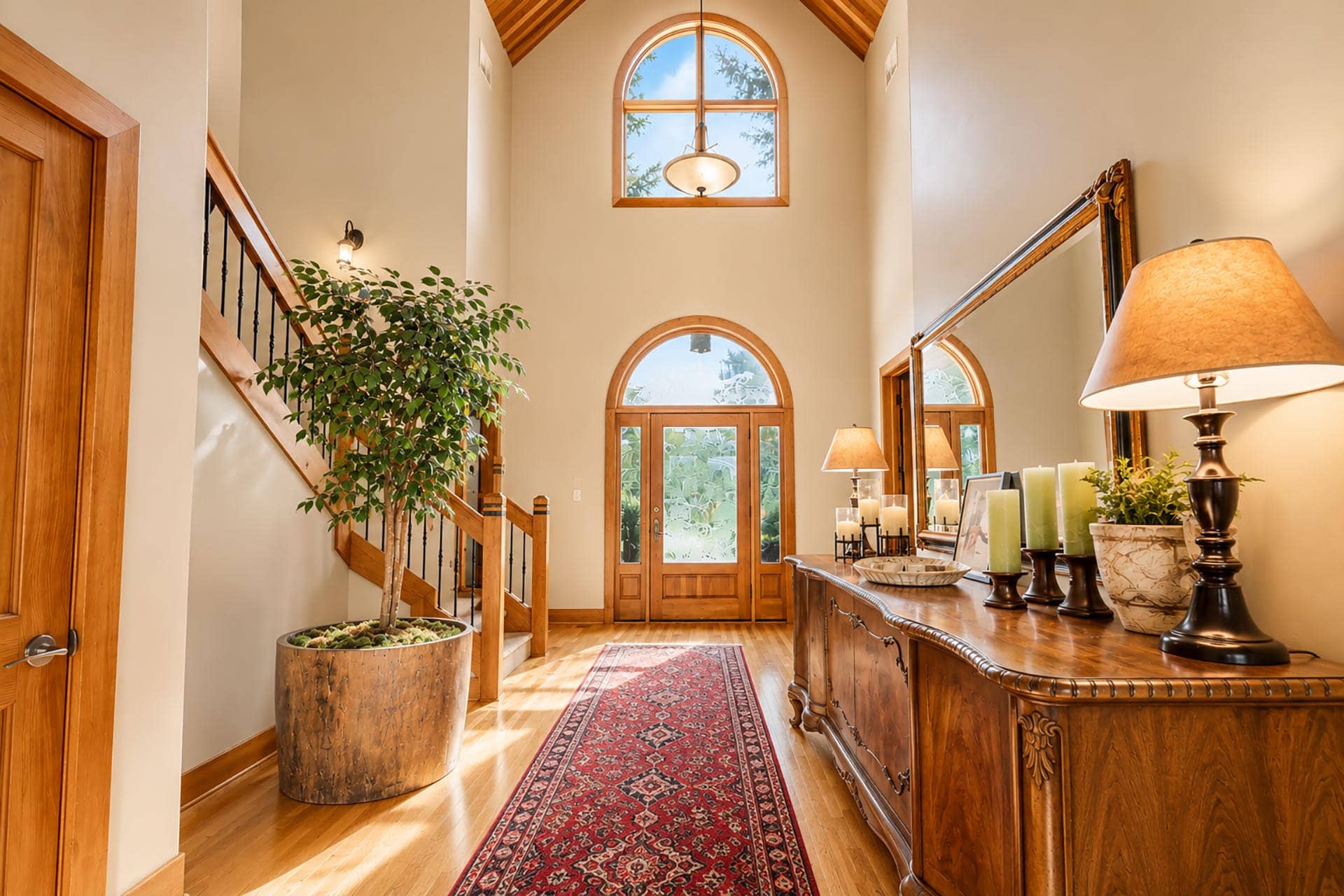 Bright foyer with a wooden staircase, tall arched windows, and a carved wooden console table holding lamps and candles.