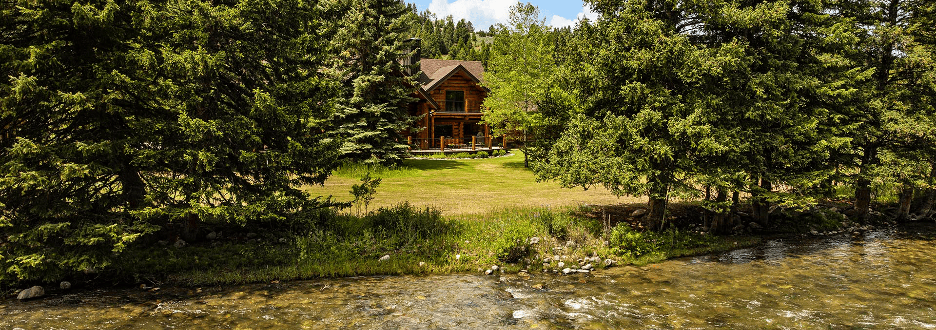 Log cabin tucked among tall evergreen trees beside a flowing river, with a grassy yard and wooden deck.