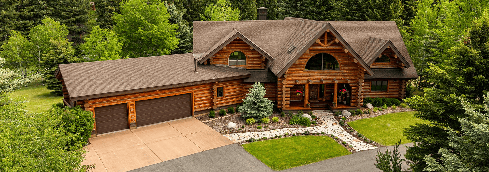 A rustic log cabin with a three-car garage set among trees, with a stone walkway and landscaped front yard.