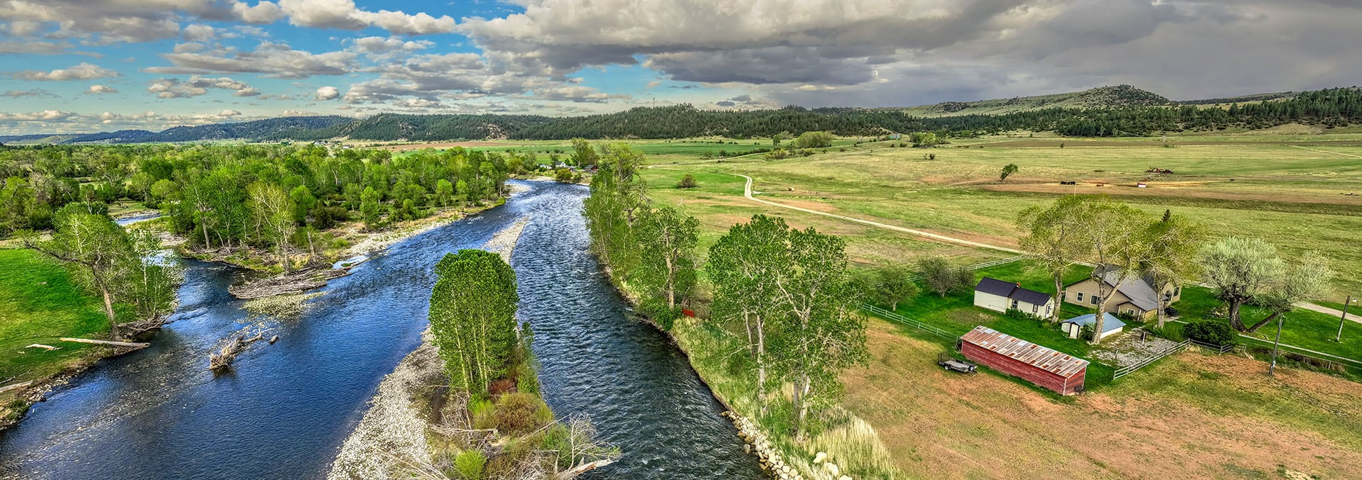 Aerial view of a winding river through a green valley, with trees along the banks and a farmstead with a red barn on the right.