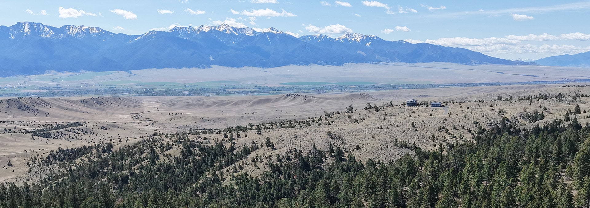 Panoramic view of a desert valley: sand dunes in the midground, evergreen forest in the foreground, and a snow-capped mountain range in the distance; a small house on a hill.