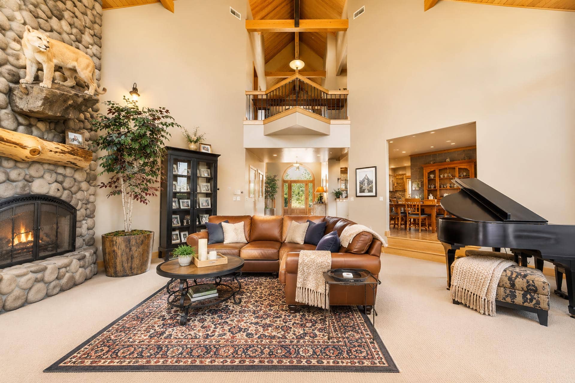 Large living room with a stone fireplace, lion statue on the mantel, leather sofa, and a grand piano along the right wall.
