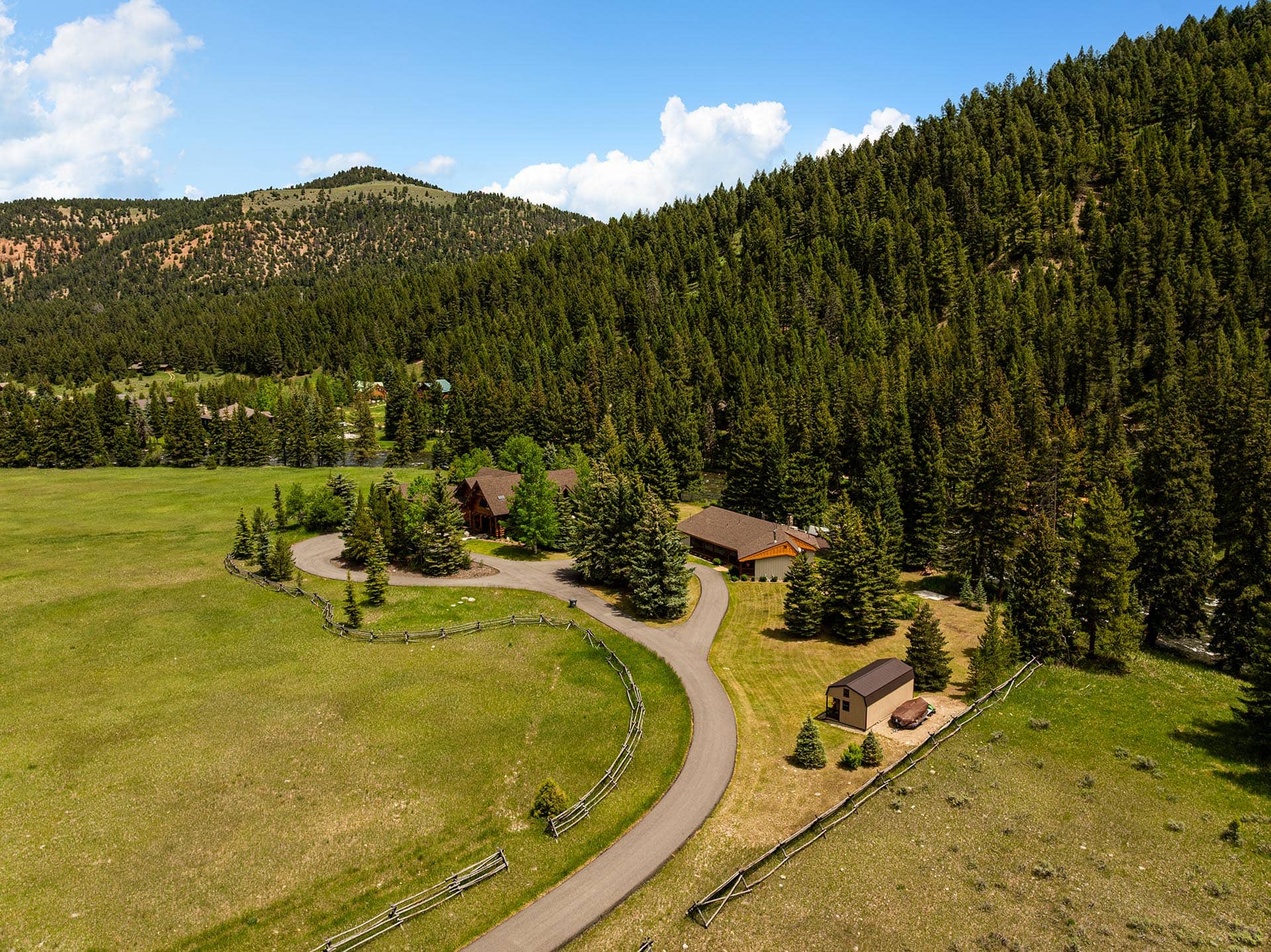 Aerial view of a rural property with a winding road, fenced green pastures, and dense pine forests against a blue sky.