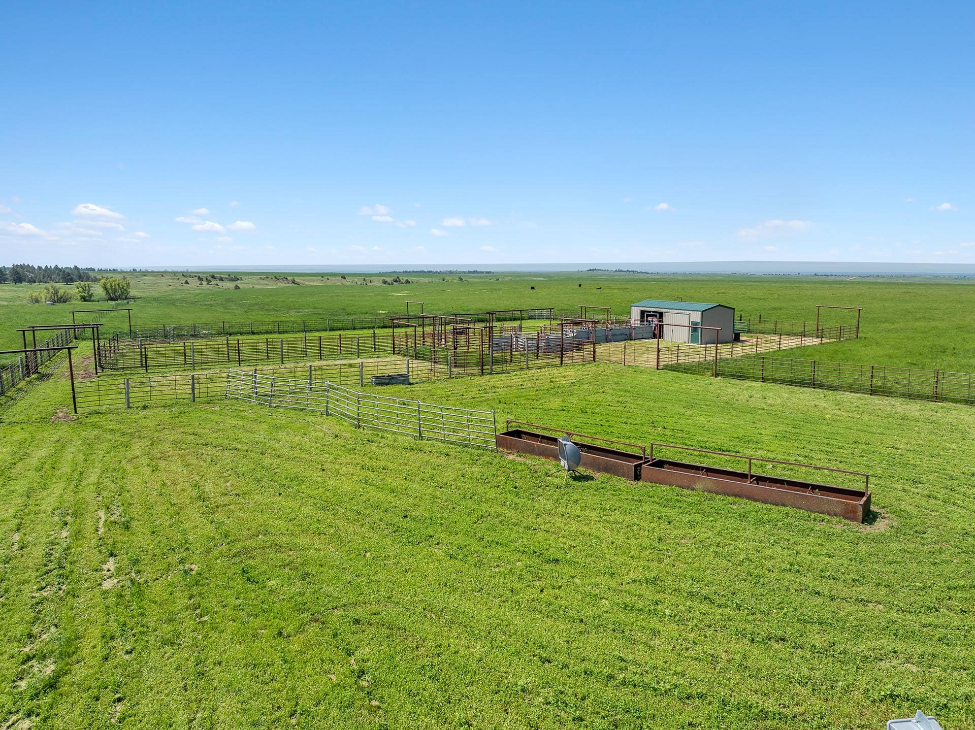 Wide view of a rural farm with multiple metal fenced pens, a small shed, and green pastures under a clear blue sky