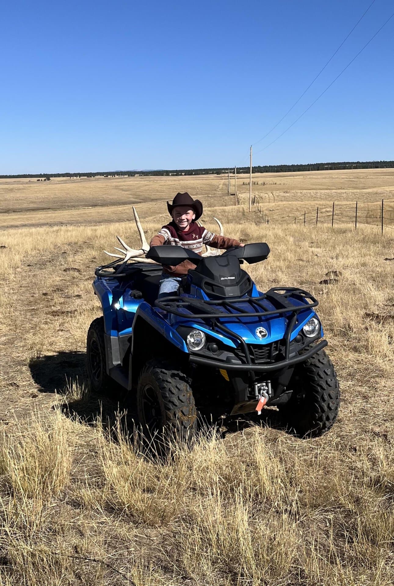 Child wearing a brown cowboy hat sits on a blue ATV in a dry, open field under a clear blue sky.