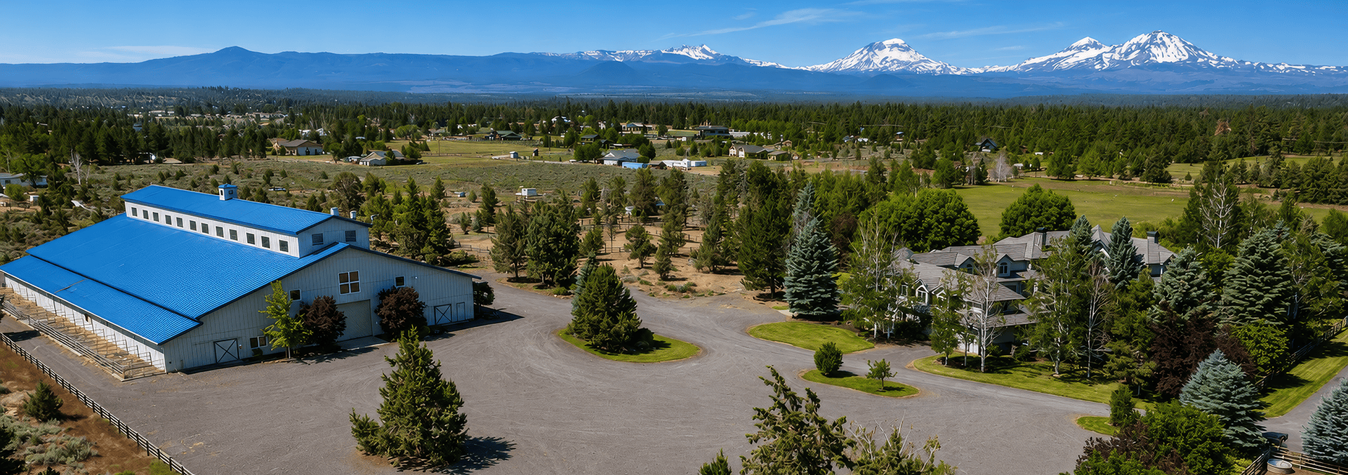 Aerial view of a rural homestead with a large blue-roof barn, circular driveway, and pine trees, with snow-capped mountains in the distance under a clear blue sky.