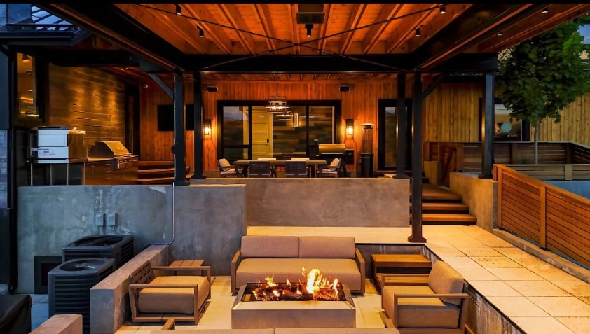 Outdoor lounge area with a rectangular fire pit surrounded by beige sectional seating under a wooden pergola, evening lighting and concrete walls nearby.