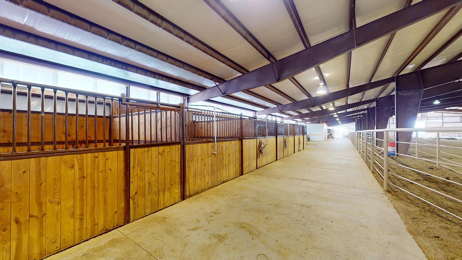 Indoor horse stable hallway with wooden stalls and metal rails, under a high ceiling with overhead lights