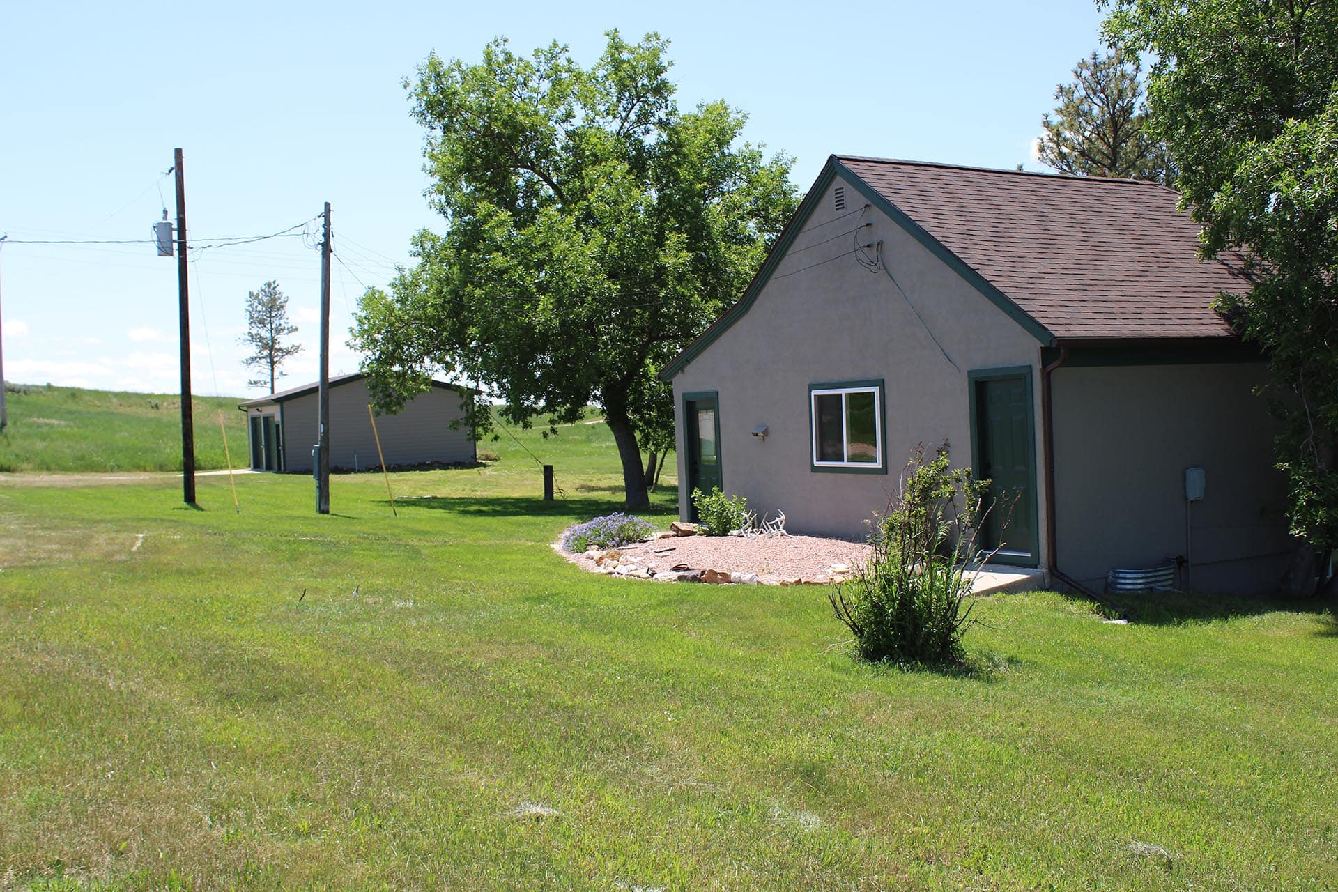 Rural scene with a beige one-story house, brown roof, and green lawn, framed by trees and power lines under a clear blue sky