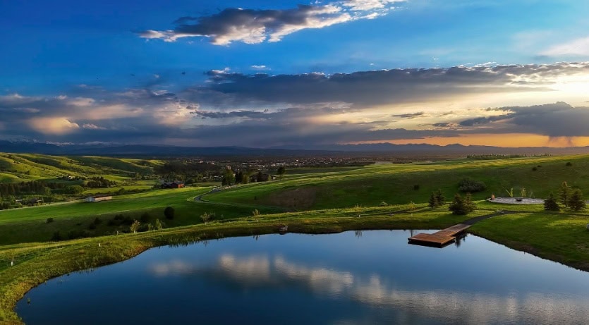 Tranquil rural landscape at sunset with a reflective pond, green rolling hills, and a wooden dock extending into the water; distant town visible on the horizon under a dramatic sky.