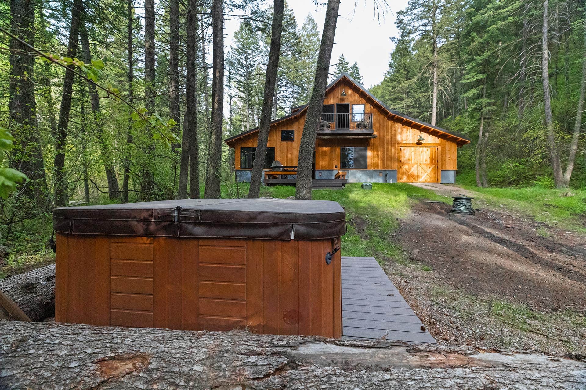 Wooden cabin surrounded by tall trees in a forest, with a hot tub in the foreground.