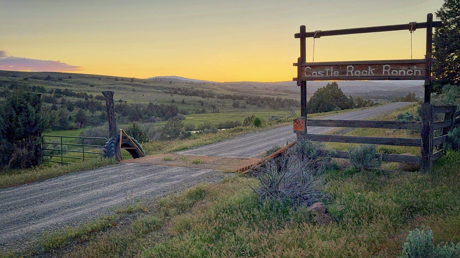 Wooden ranch gate with 'Castle Rock Ranch' sign at sunset over a wide, grassy valley road.