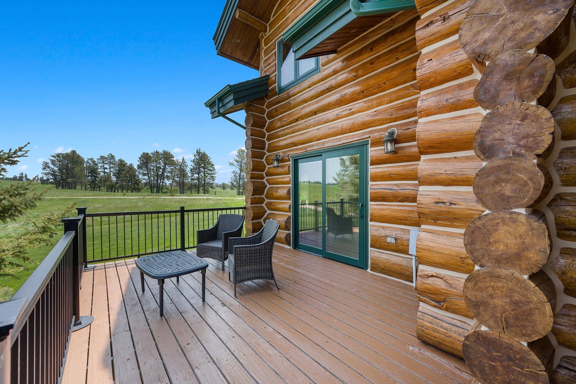 Deck of a log cabin with wicker seating and a coffee table, sliding glass door, and stacked log ends, overlooking a grassy field and blue sky.