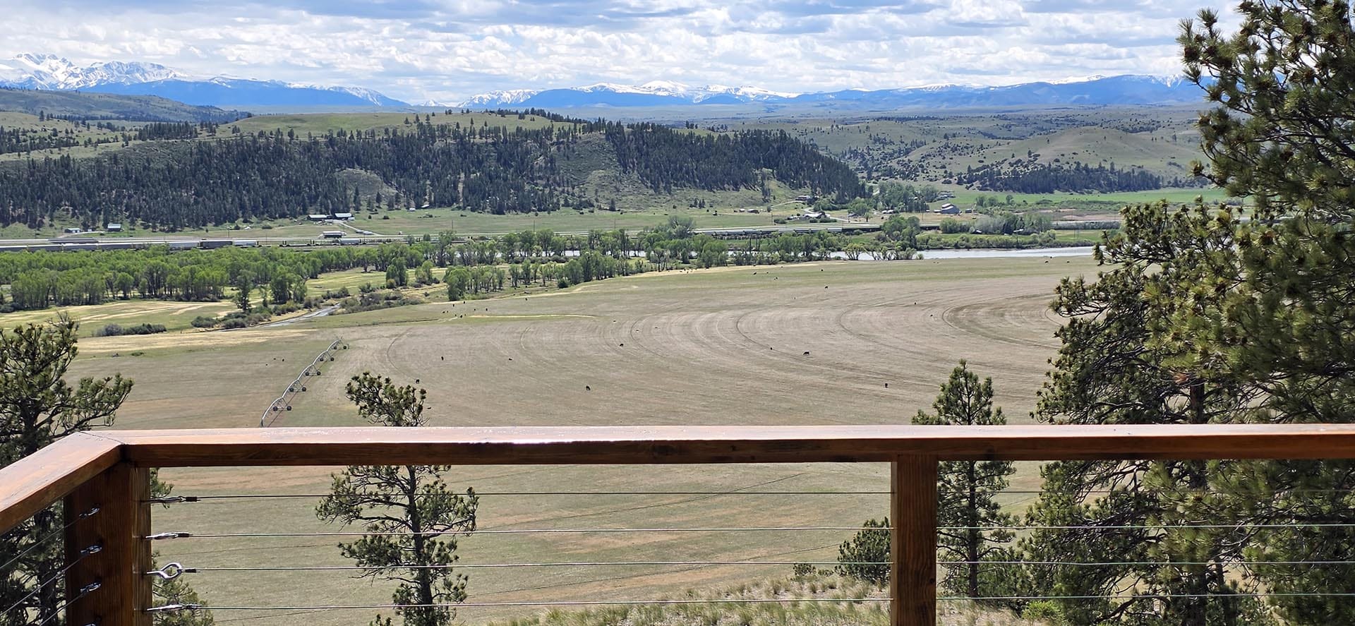 Panoramic valley scene with green fields, scattered trees, and distant snow‑capped mountains, seen from a wooden deck railing in the foreground.