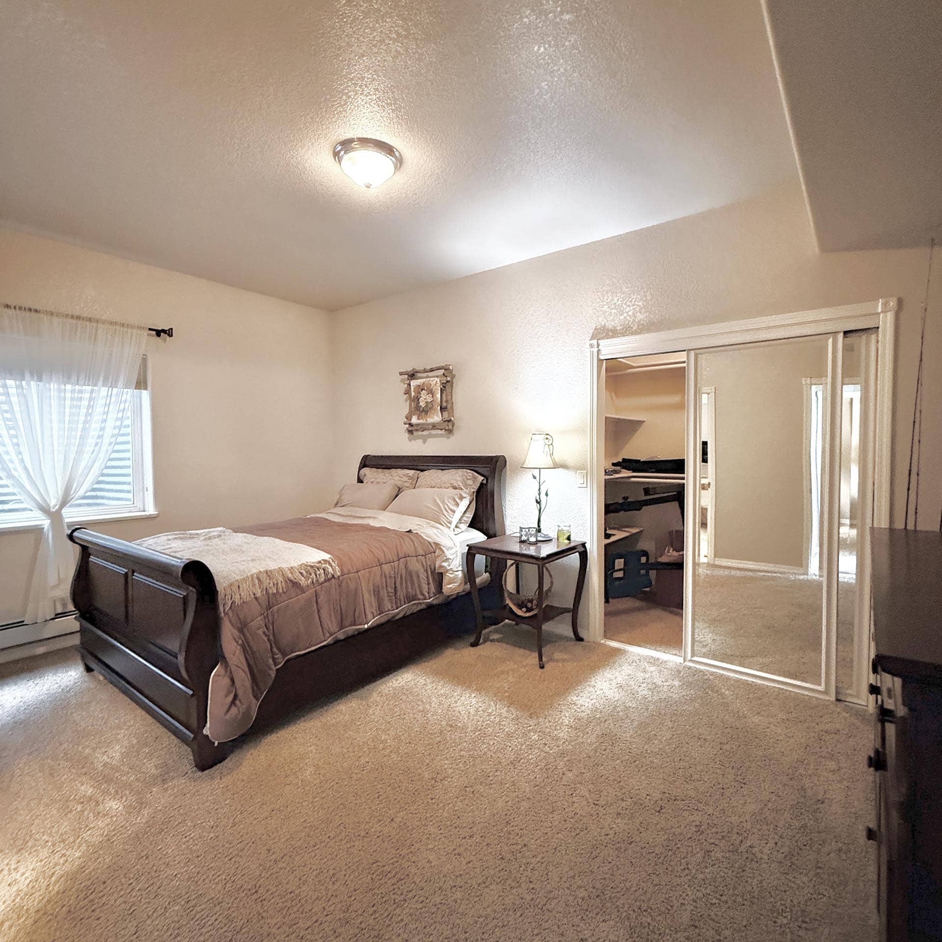 Bedroom with a bed, nightstand, lamp, and mirrored dresser; beige walls and a window with sheer curtains on the left