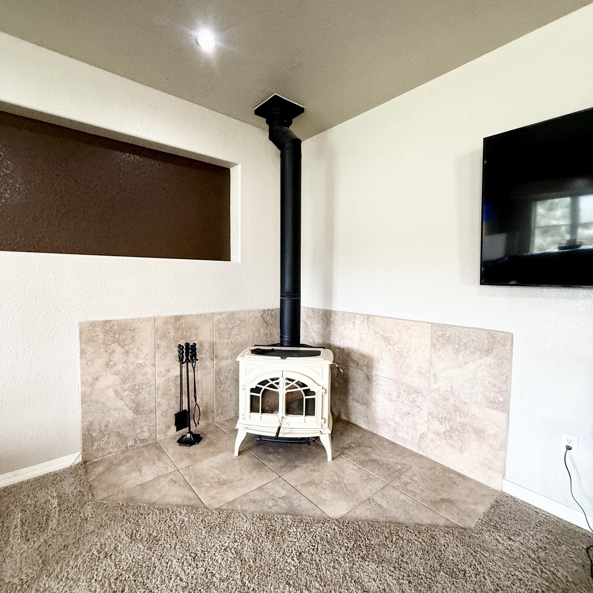 White wood stove with a black chimney pipe in a tiled corner, TV mounted on the wall nearby, and beige carpeting.