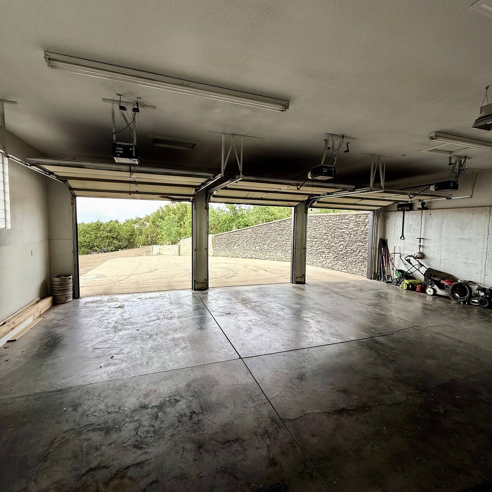 Empty garage with open doors and scattered lawn equipment along the right wall, concrete floor visible outside greenery.