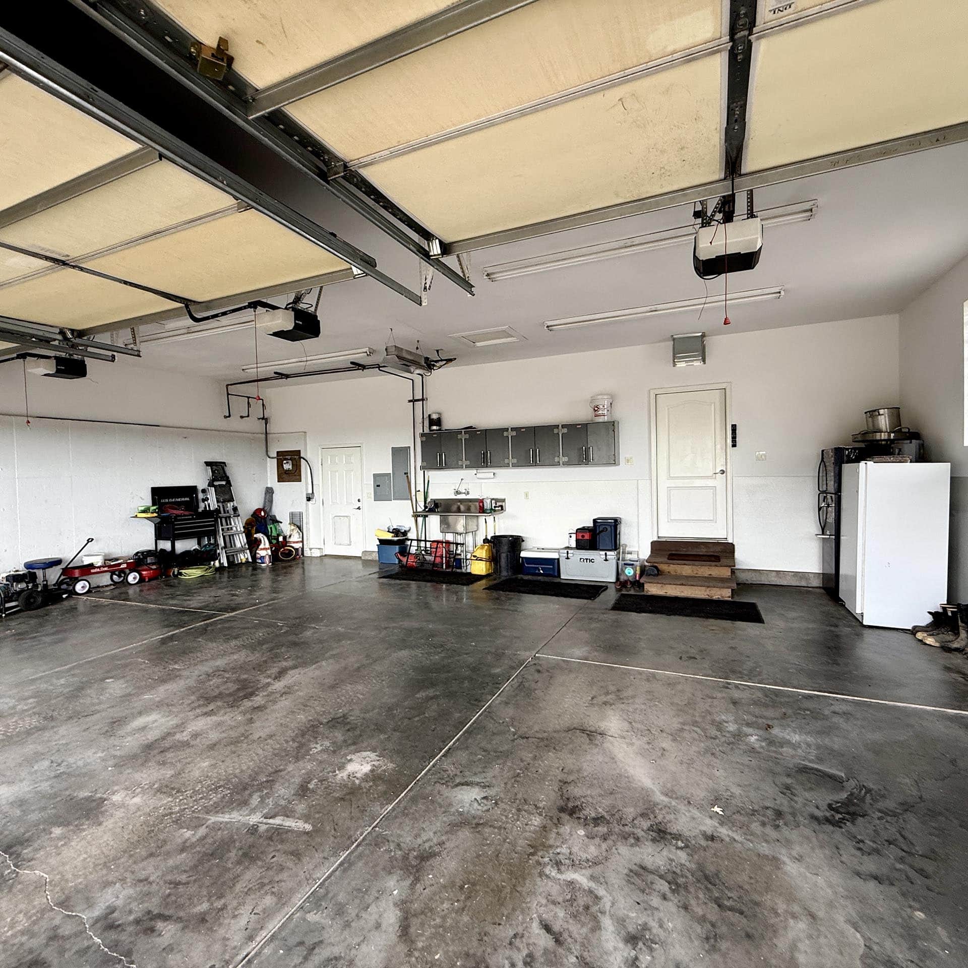 Spacious garage interior with overhead door tracks, cluttered workbench, and storage cabinets along the back wall; concrete floor with stains and tools scattered around.