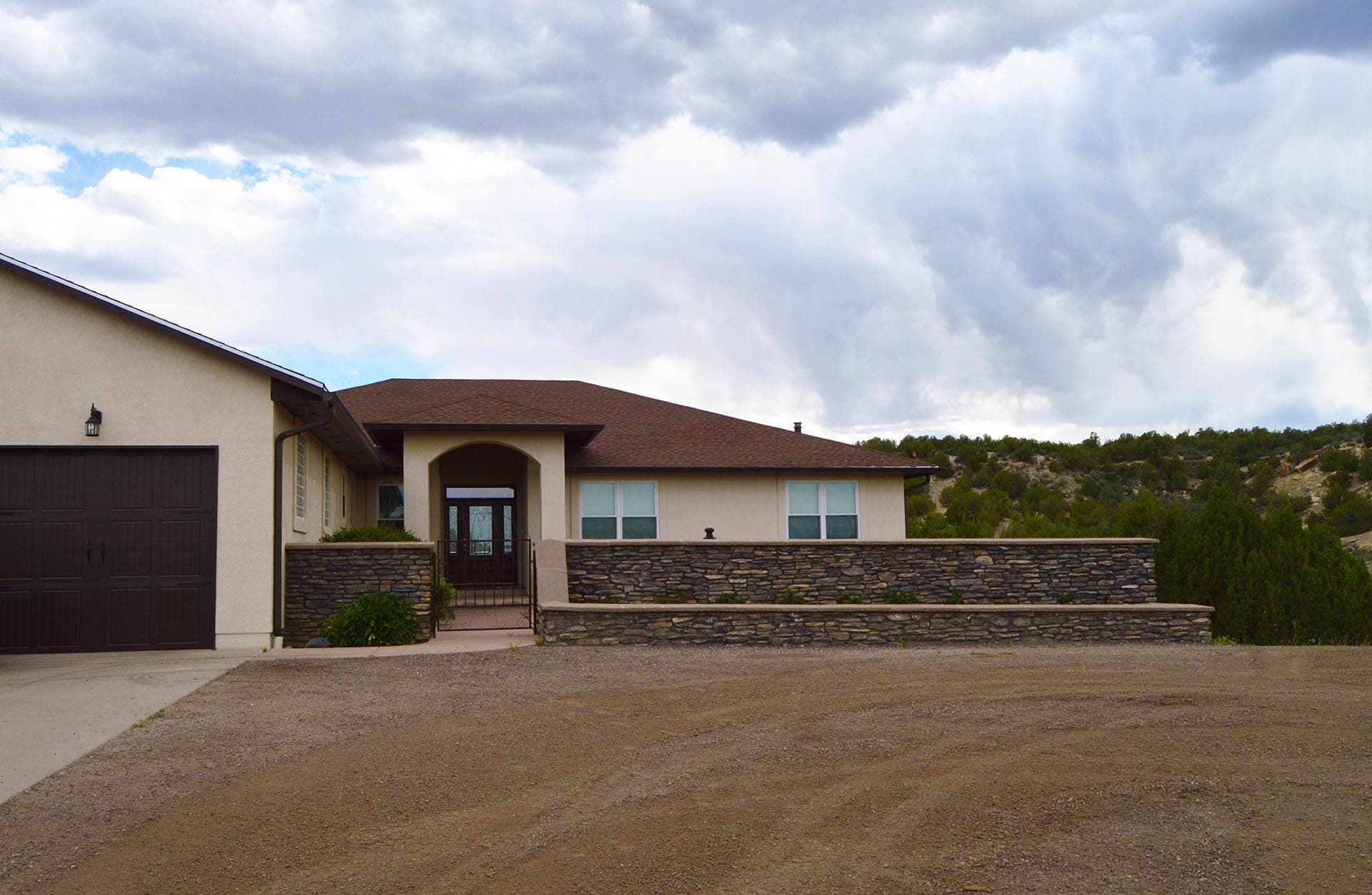 Front view of a beige modern house with a dark garage door, stone accents, and a dirt driveway beneath a cloudy sky.