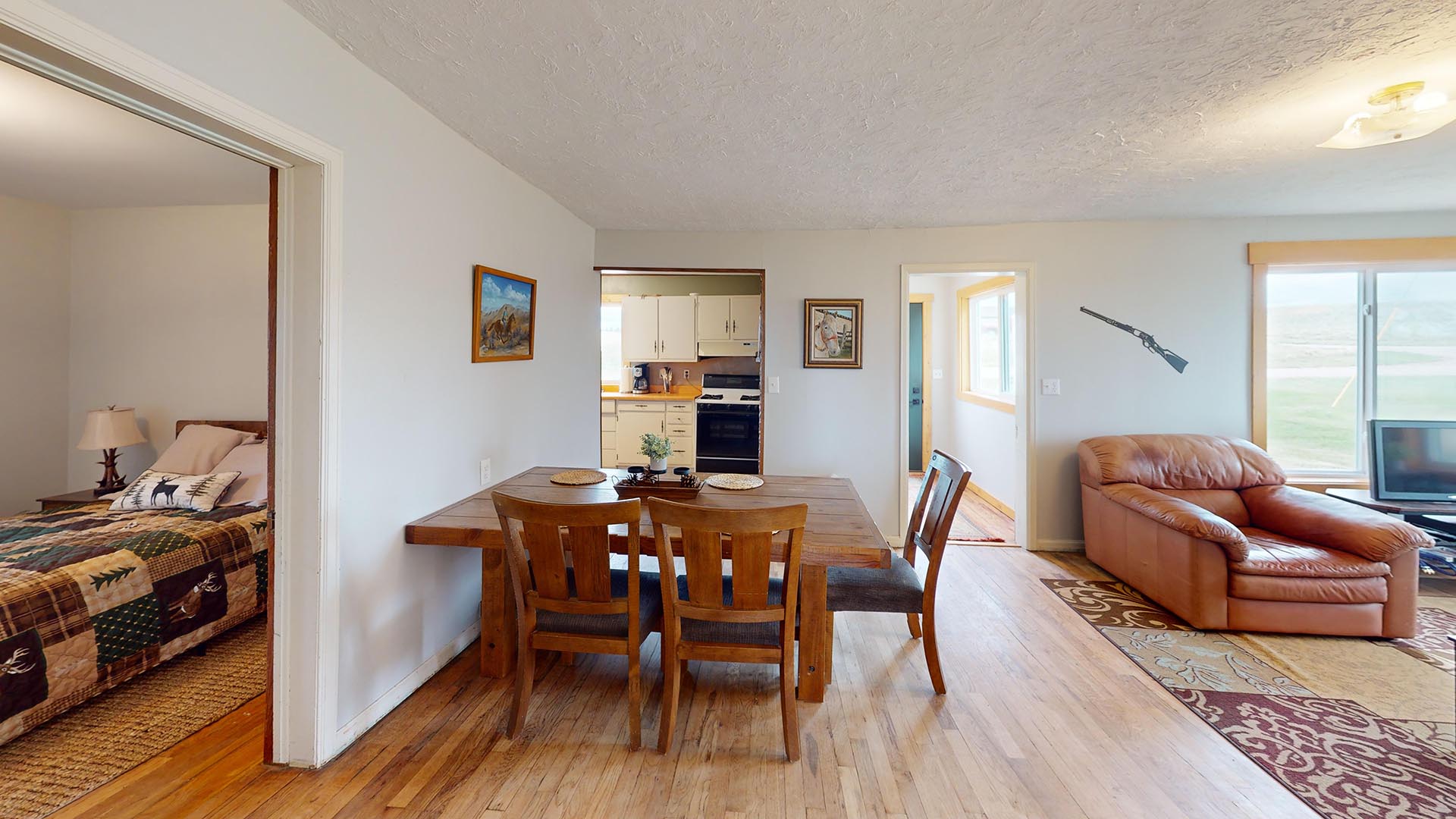 Open-plan dining and living room with a wooden table and four chairs, a tan leather sofa by a large window, and a doorway to a small kitchen; a bedroom is visible to the left through an adjacent doorway.