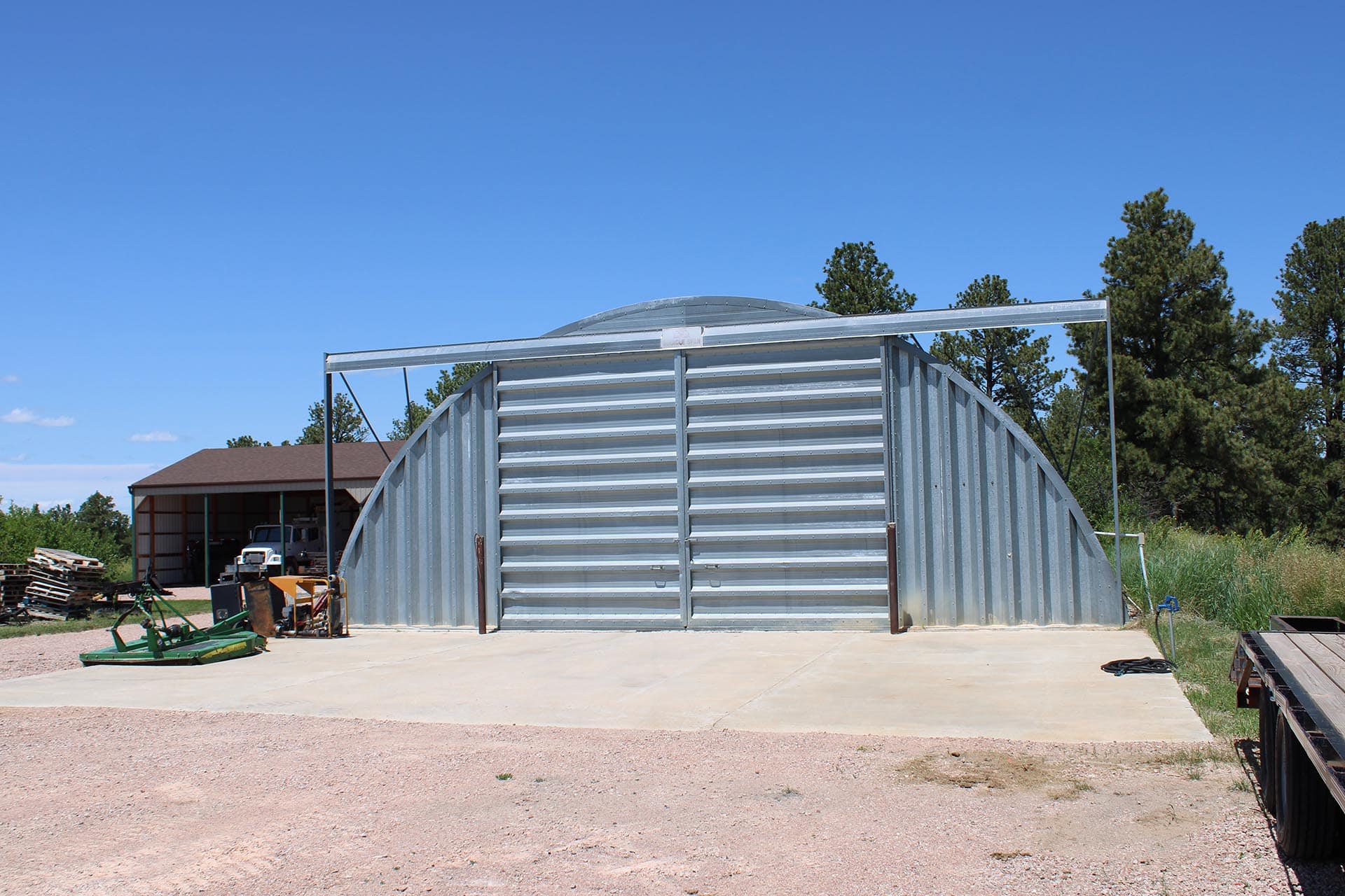 Curved corrugated metal storage shed in a rural yard with blue sky and trees behind.