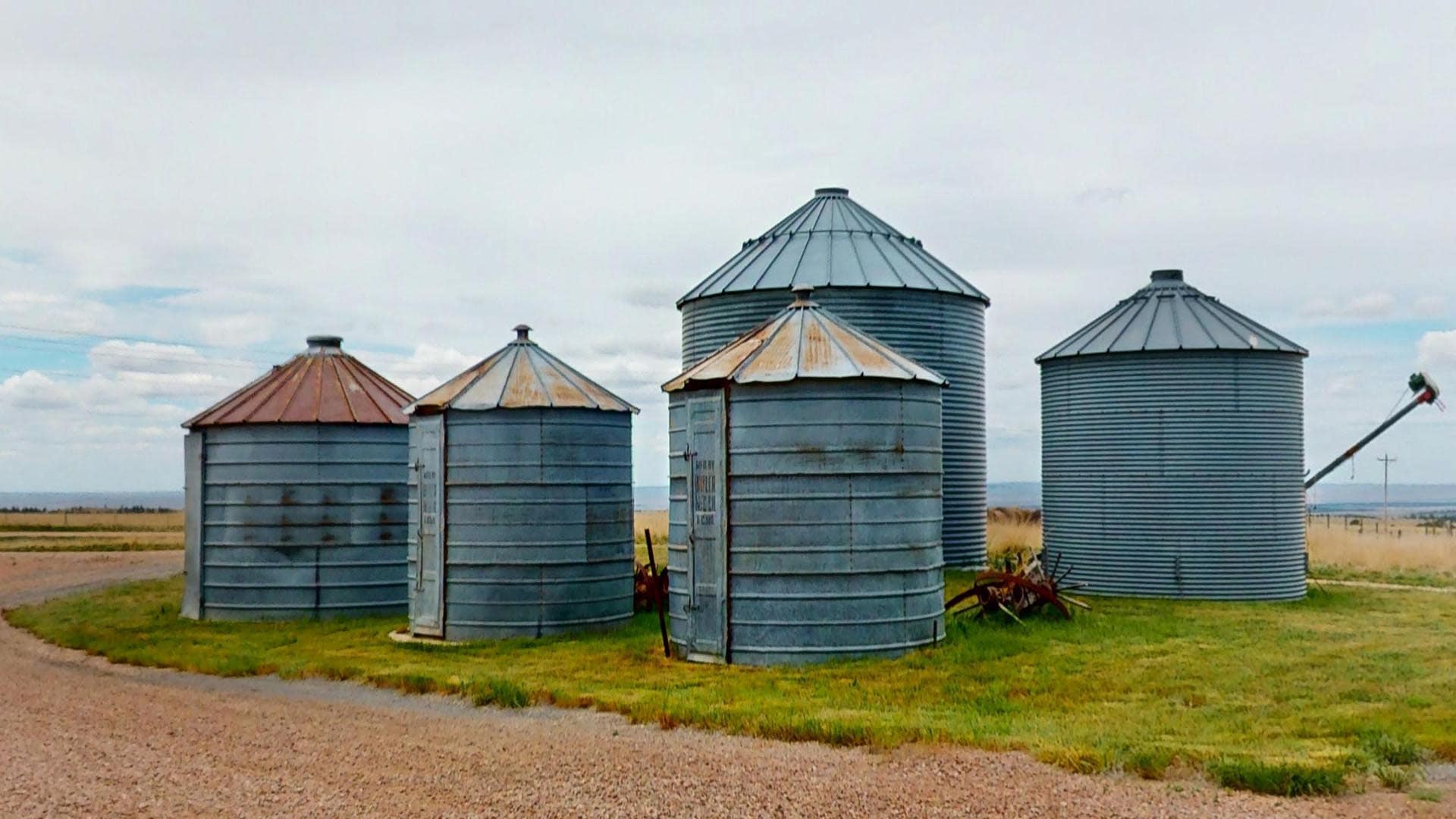 Group of metal grain silos with rusted conical roofs on a flat rural landscape.