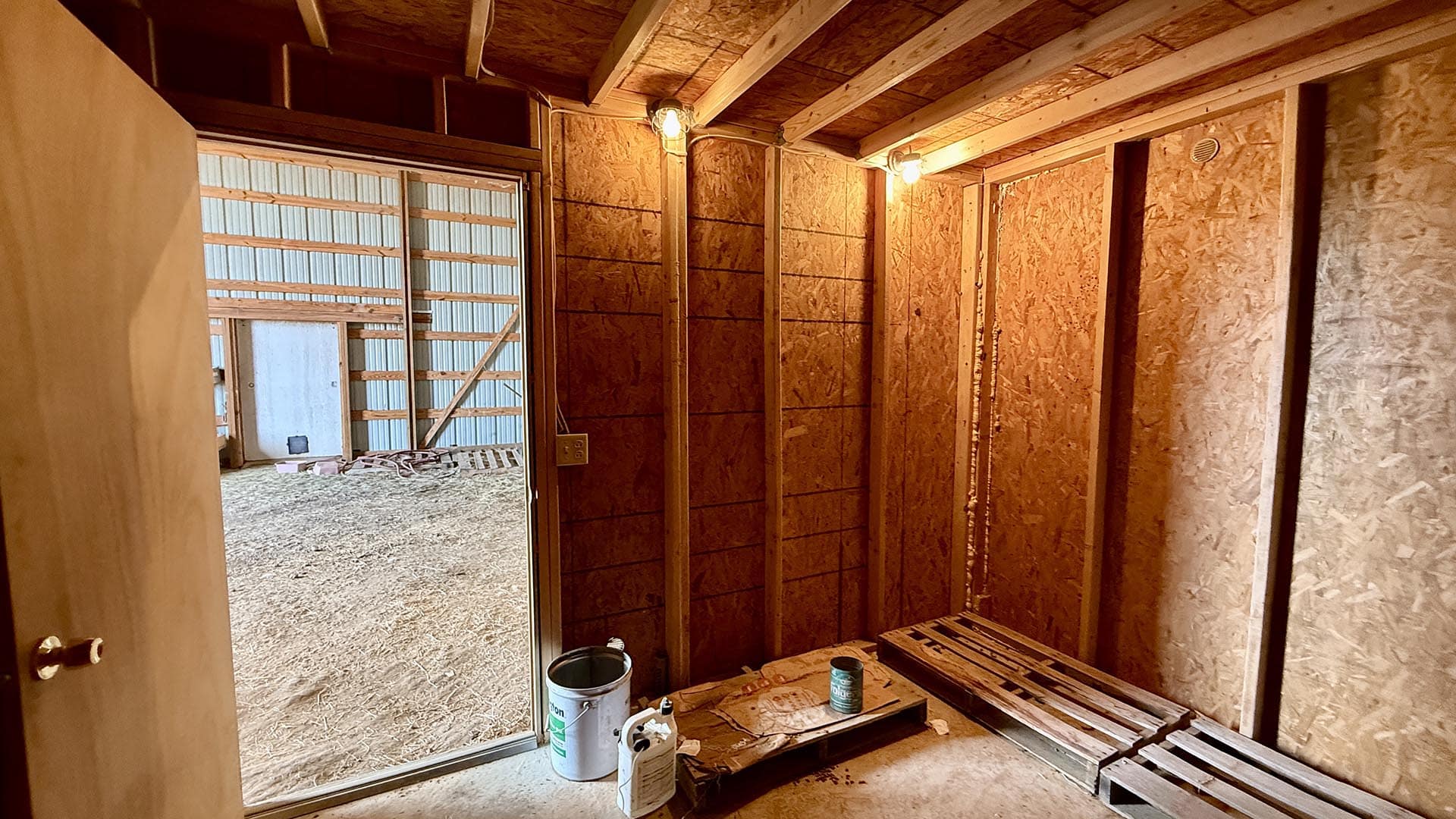Construction site interior with framed walls, plywood sheathing, and exposed ceiling joists; tools and paint cans on the floor nearby.