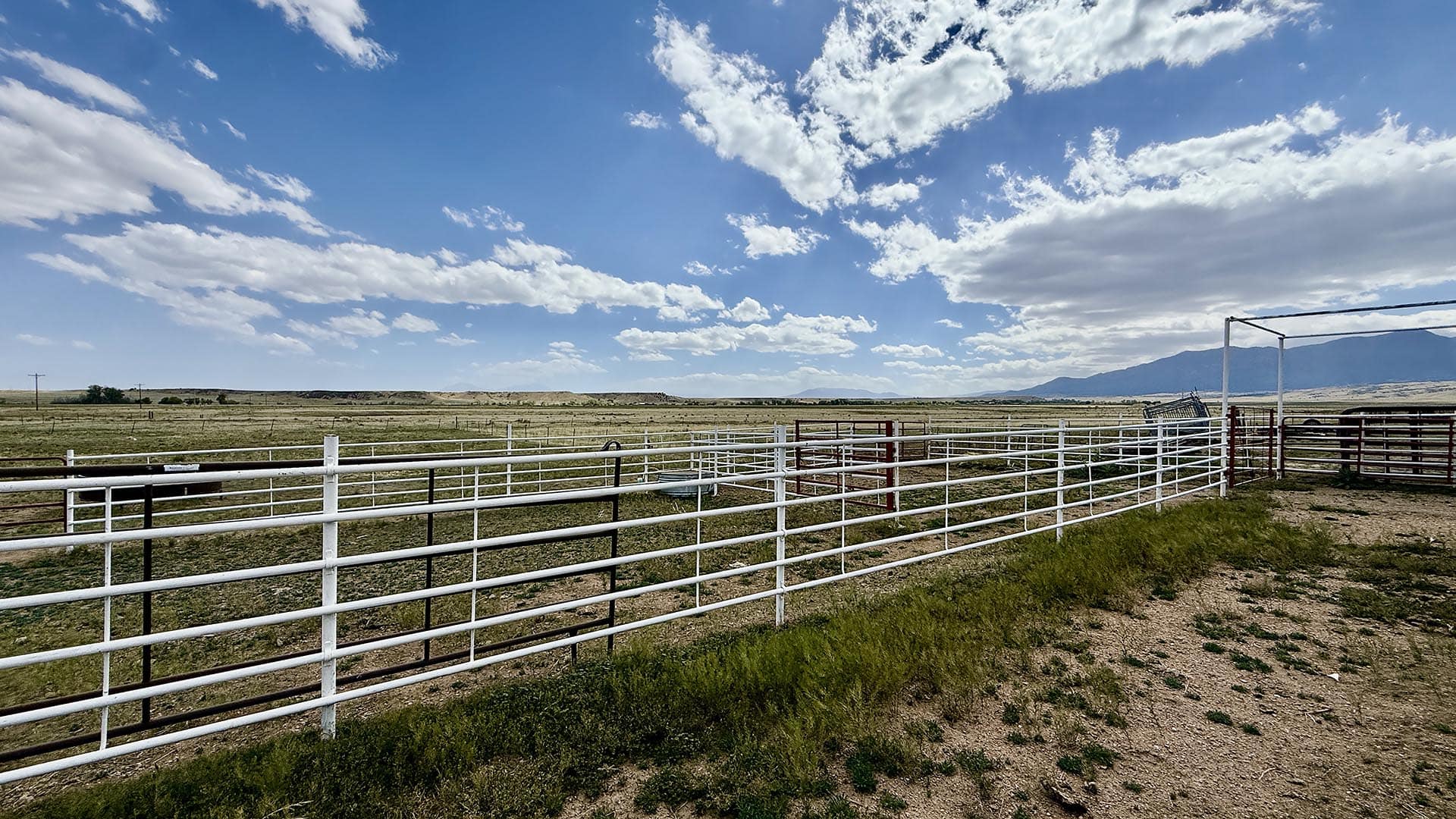 Wide view of a ranch with white metal fences, open pasture, and distant mountains under a blue sky with clouds.