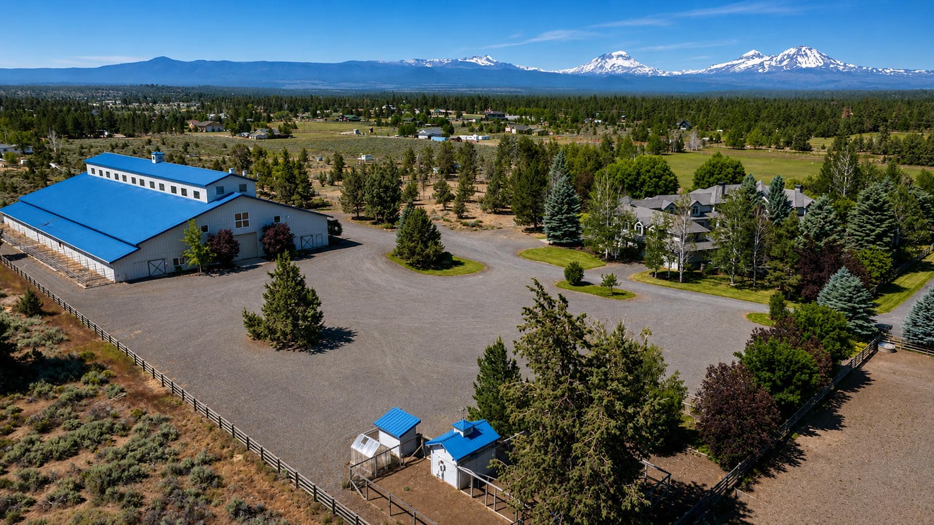 Aerial view of a rural farm with a large blue-roof barn, gravel lots, and surrounding houses, set against snow-capped mountains in the distance.