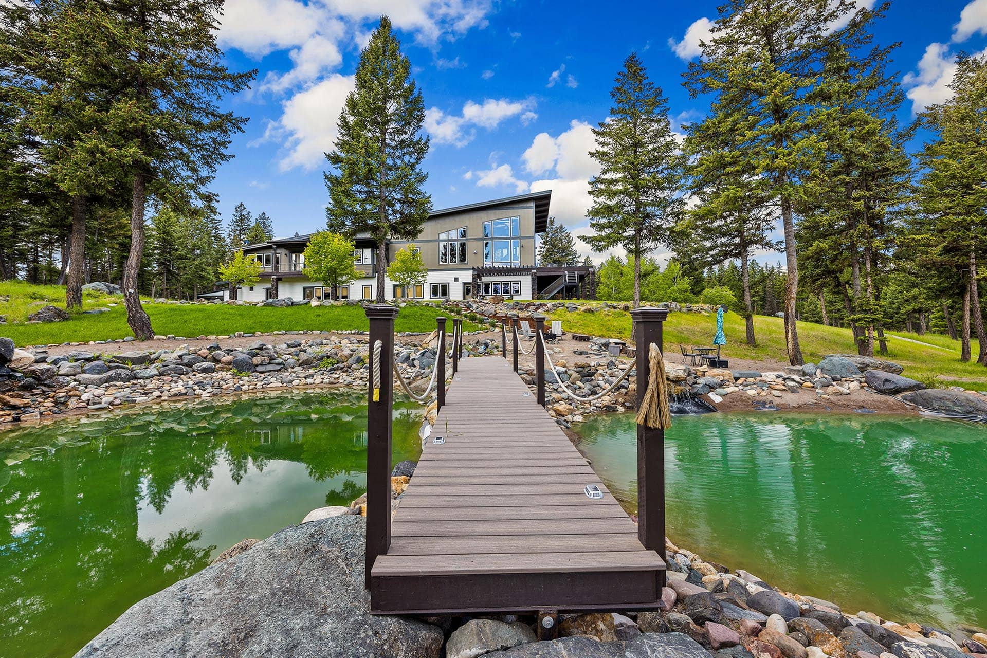 Contemporary house on a grassy lakeshore, with a wooden dock extending toward the green water and surrounding pine trees.