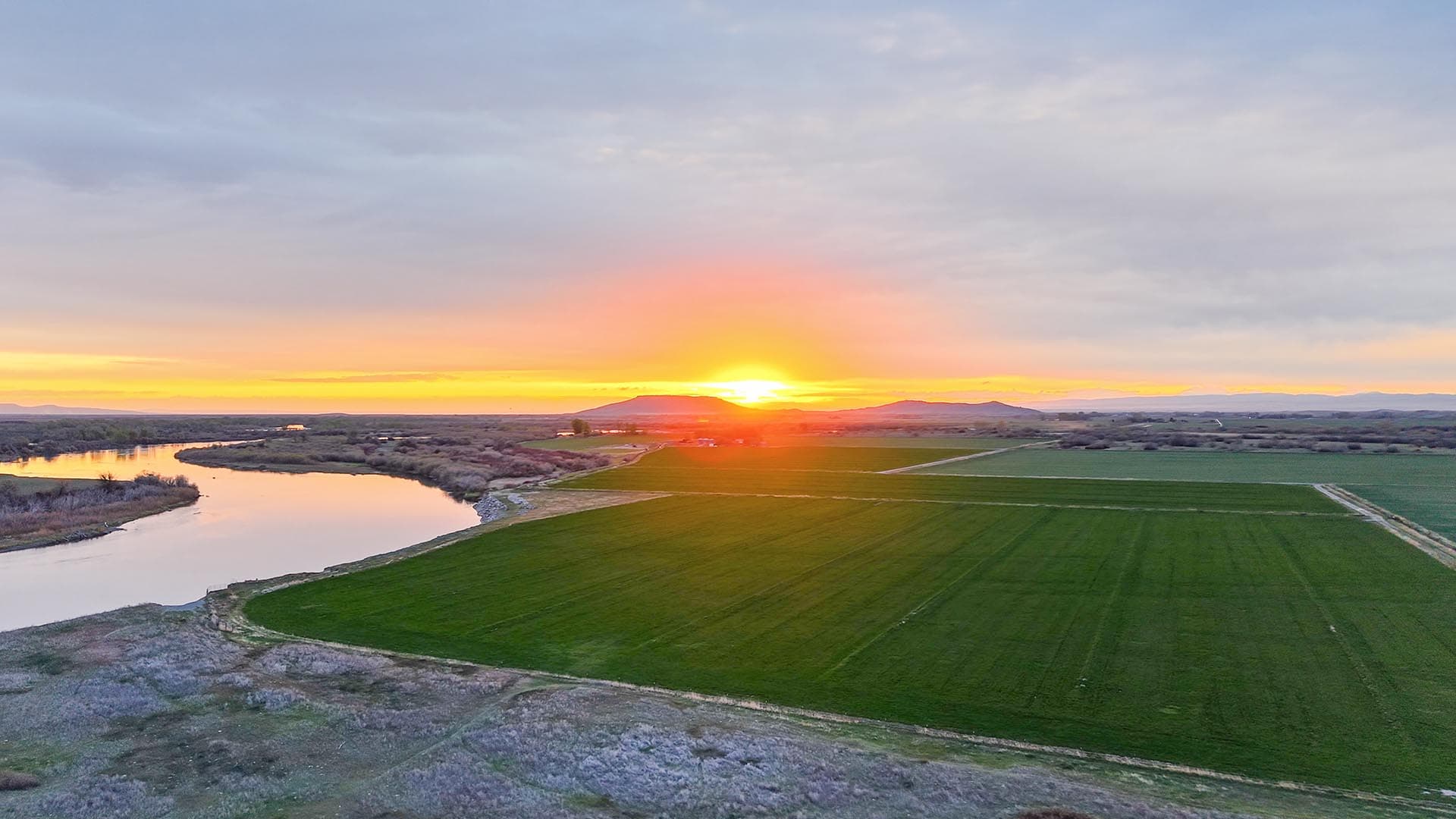 Sunset over green fields with a winding river on the left and distant hills on the horizon