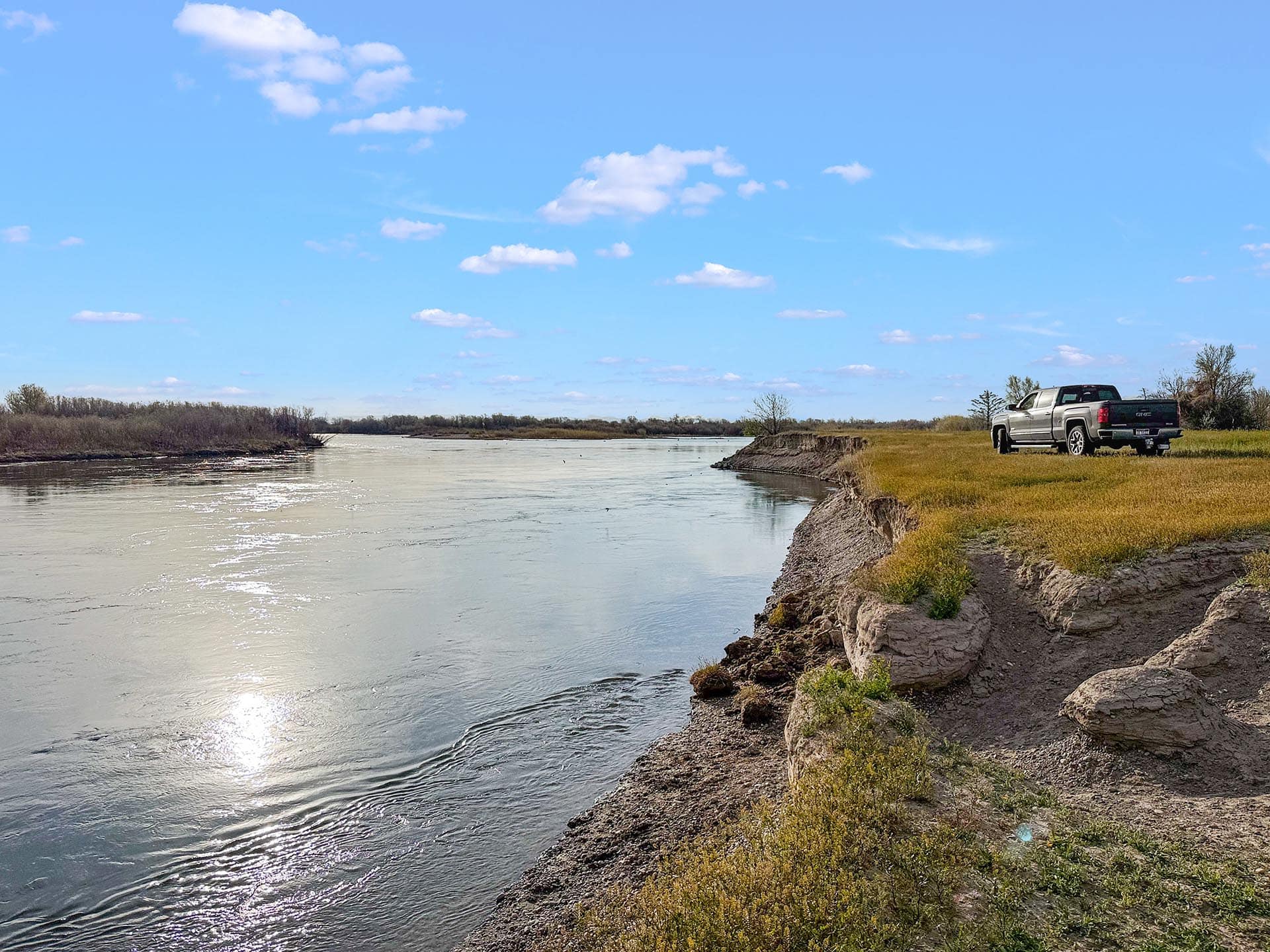 Silver pickup truck parked on a grassy riverbank with a wide river and blue sky.