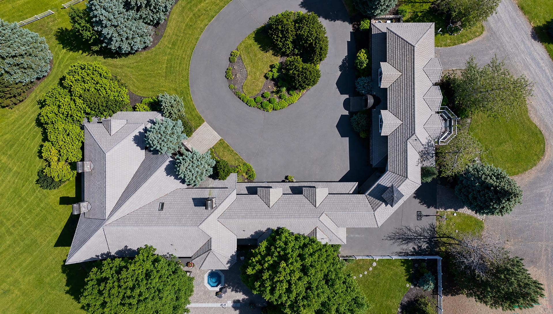 Aerial view of a large gray-roofed house with a circular driveway, manicured lawn, and surrounding trees.